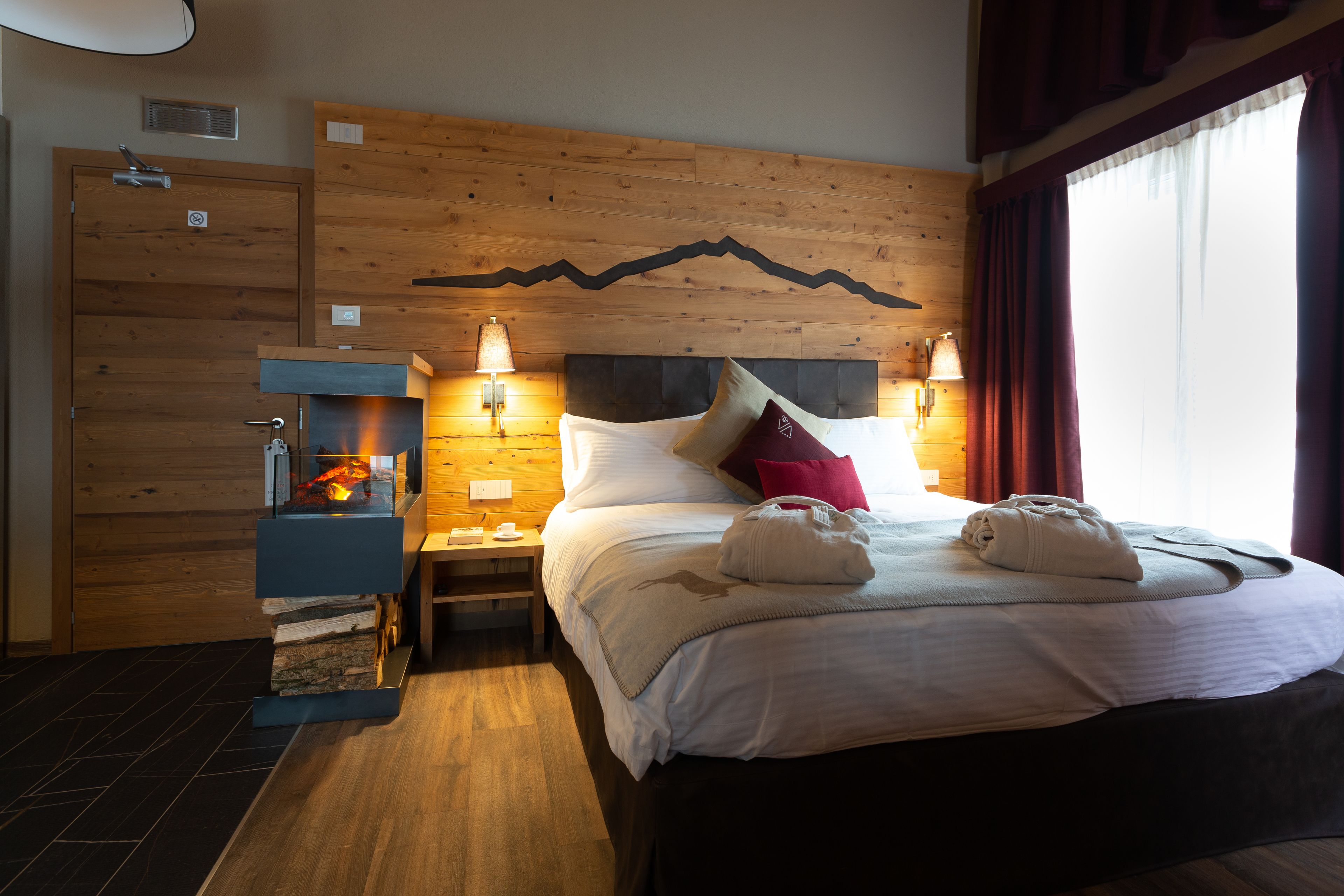 Habitación de hotel con cabecera de madera, cama con ropa blanca, dos lámparas de noche, mesita de noche y chimenea empotrada.