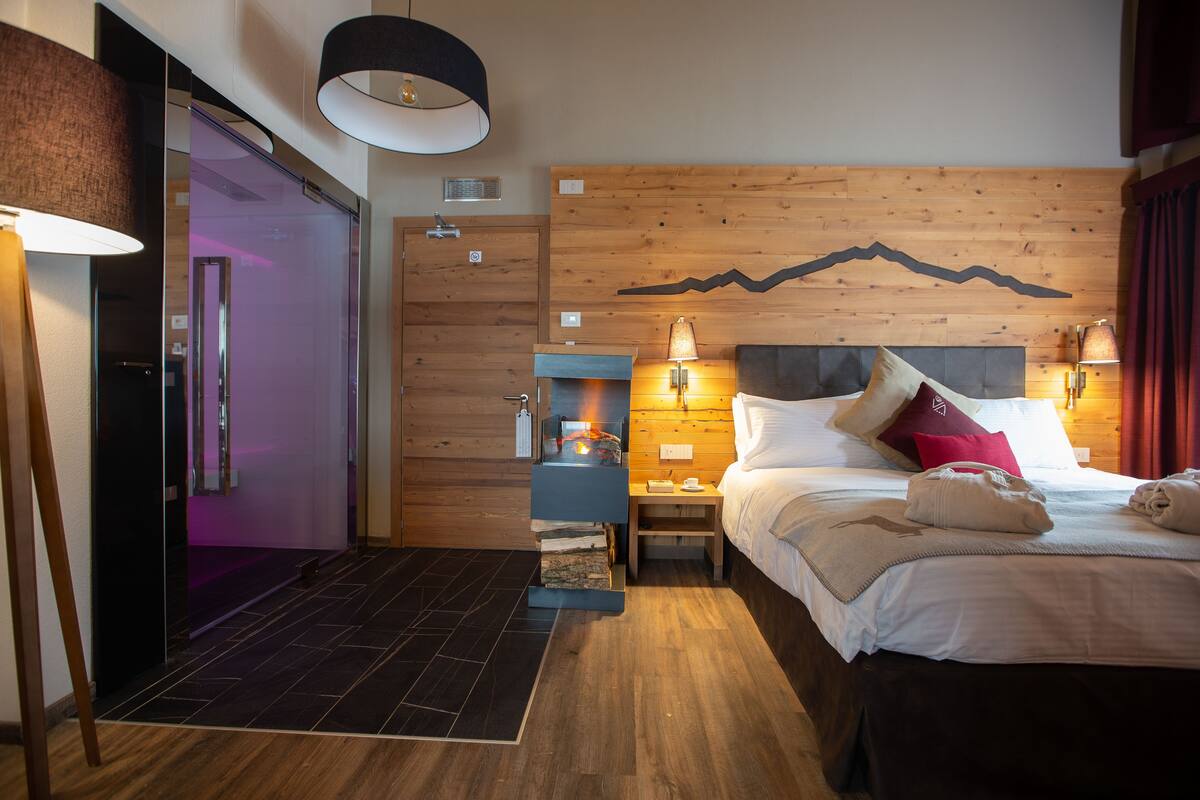 Una habitación de hotel moderna con un cabecero de madera, una cama con sábanas blancas, una mesita de noche con lámpara y una chimenea.