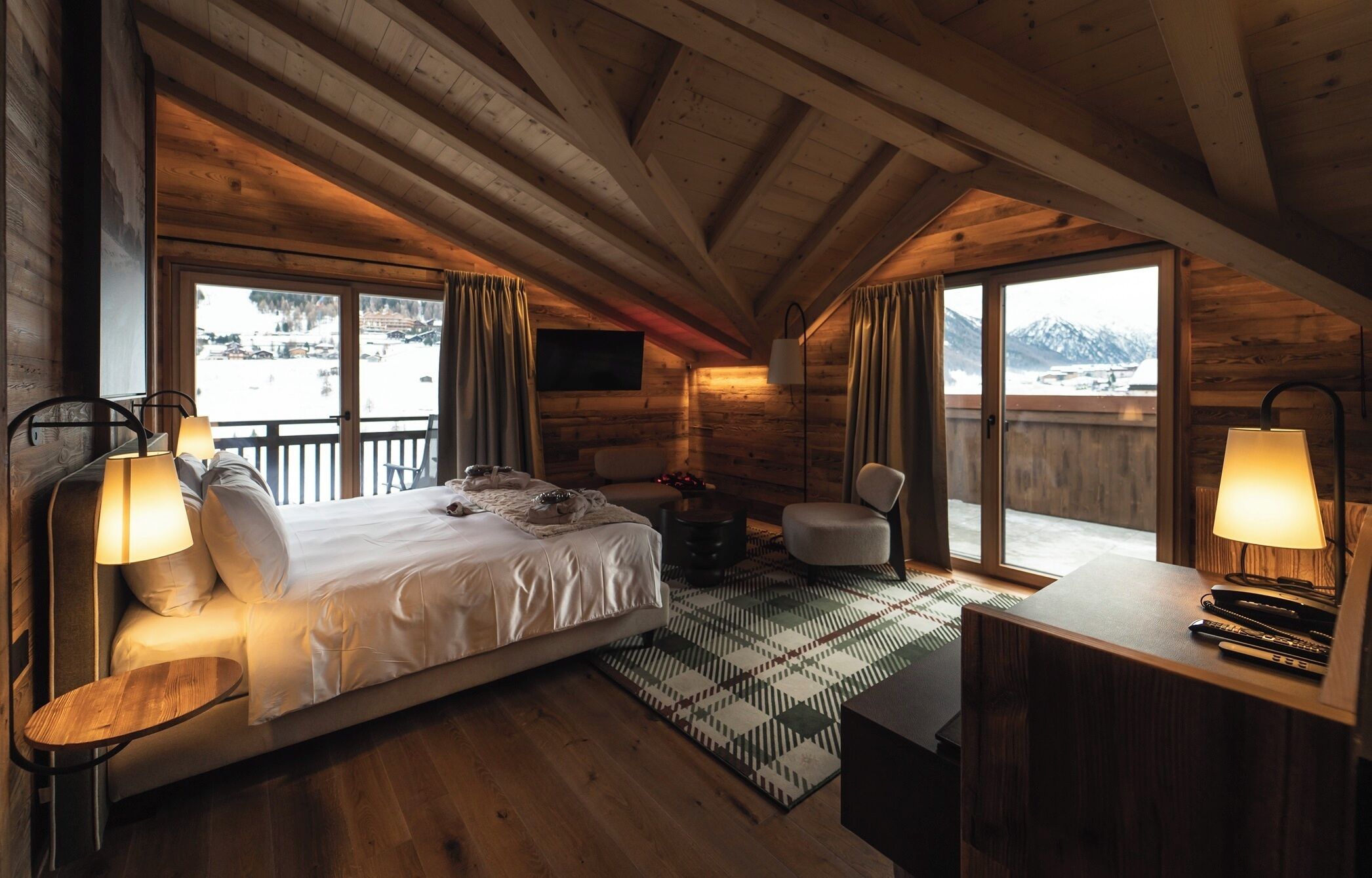 Ein gemütliches Zimmer mit Bett, Schreibtisch und Stuhl, großen Fenstern mit Bergblick und Holzwänden.