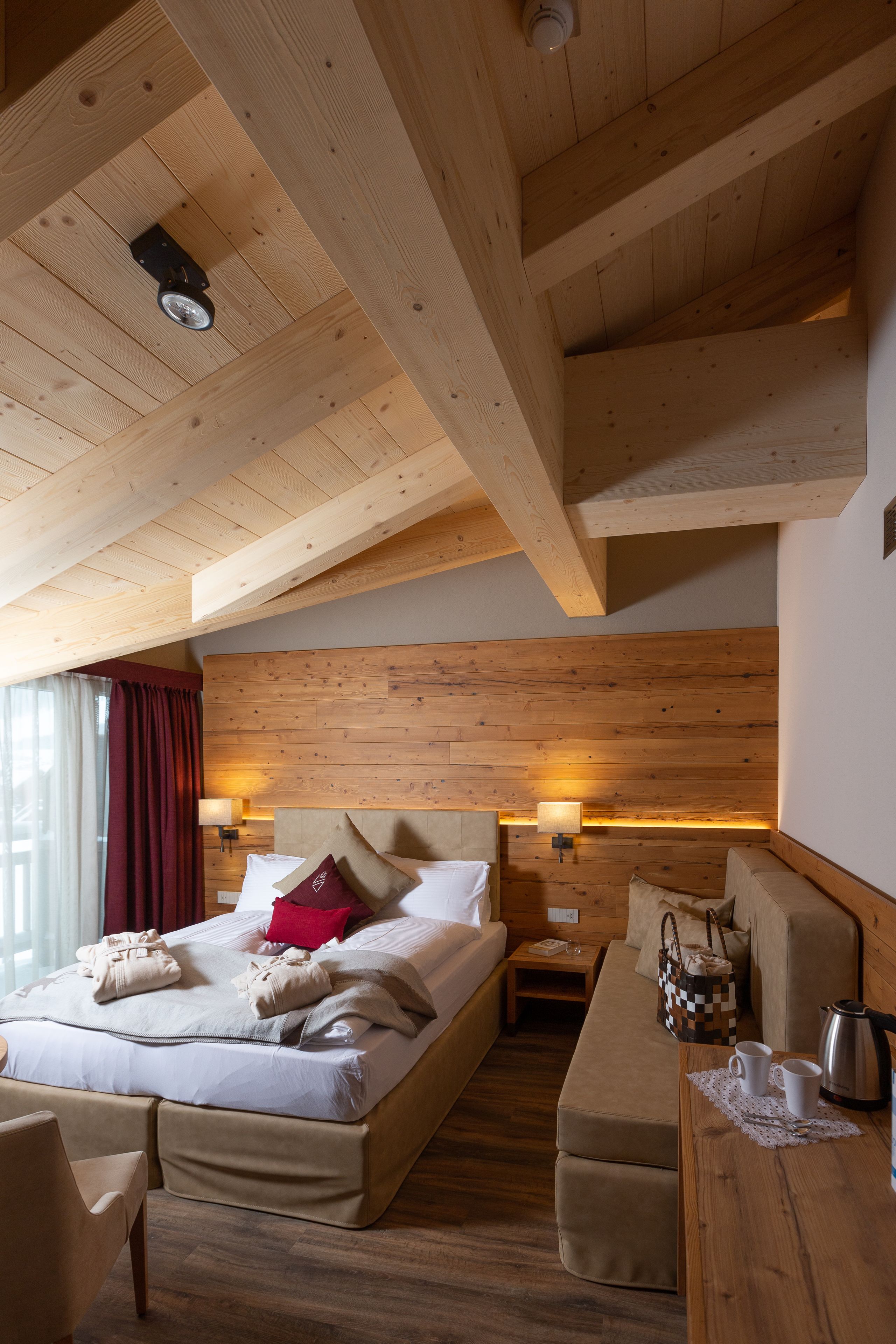 A hotel room with a wooden ceiling, a large bed with pillows, a bedside table, a sofa, and a wooden floor.