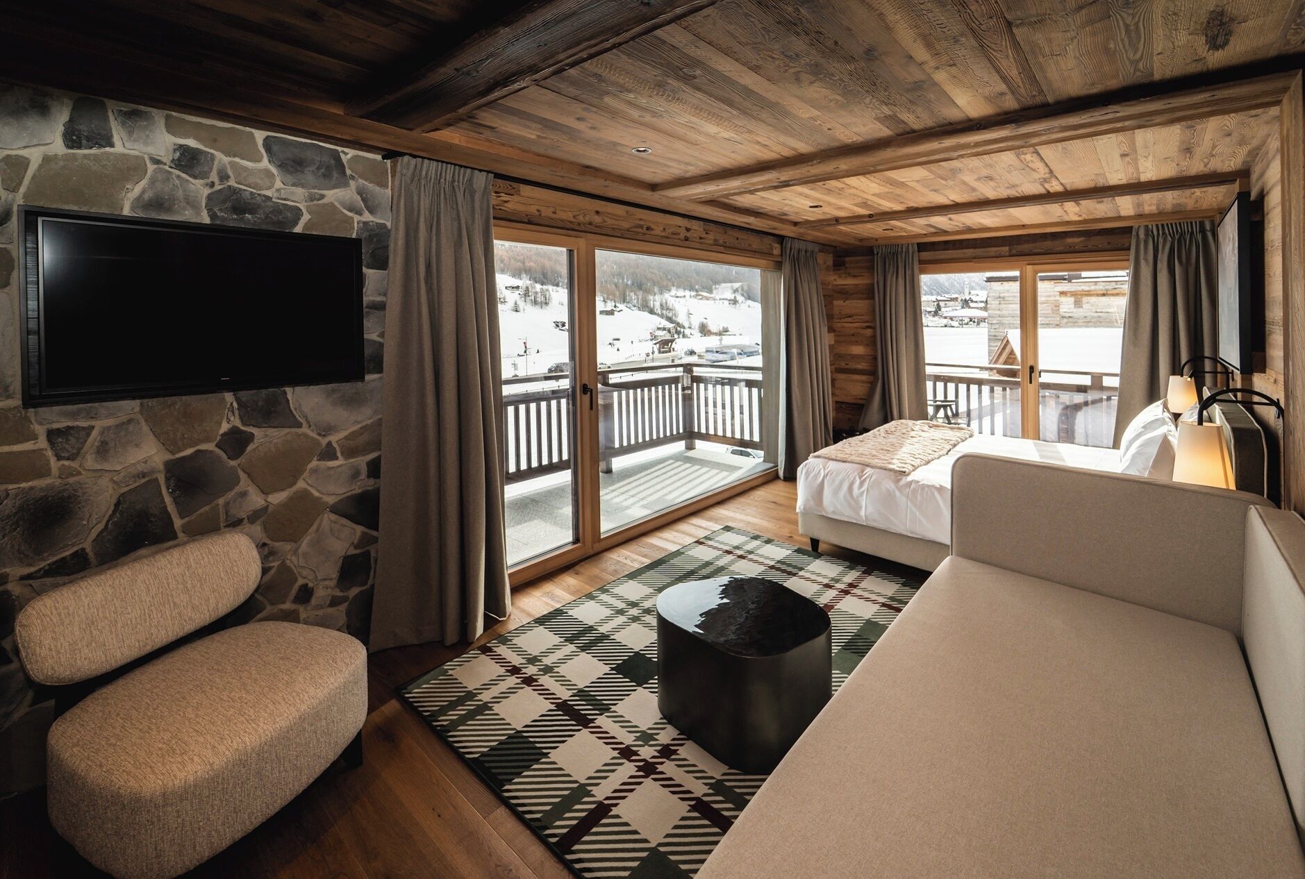 Een gezellige kamer met een stenen muur, een flatscreen televisie, een bank, een bed en een balkon met uitzicht op een besneeerd landschap.