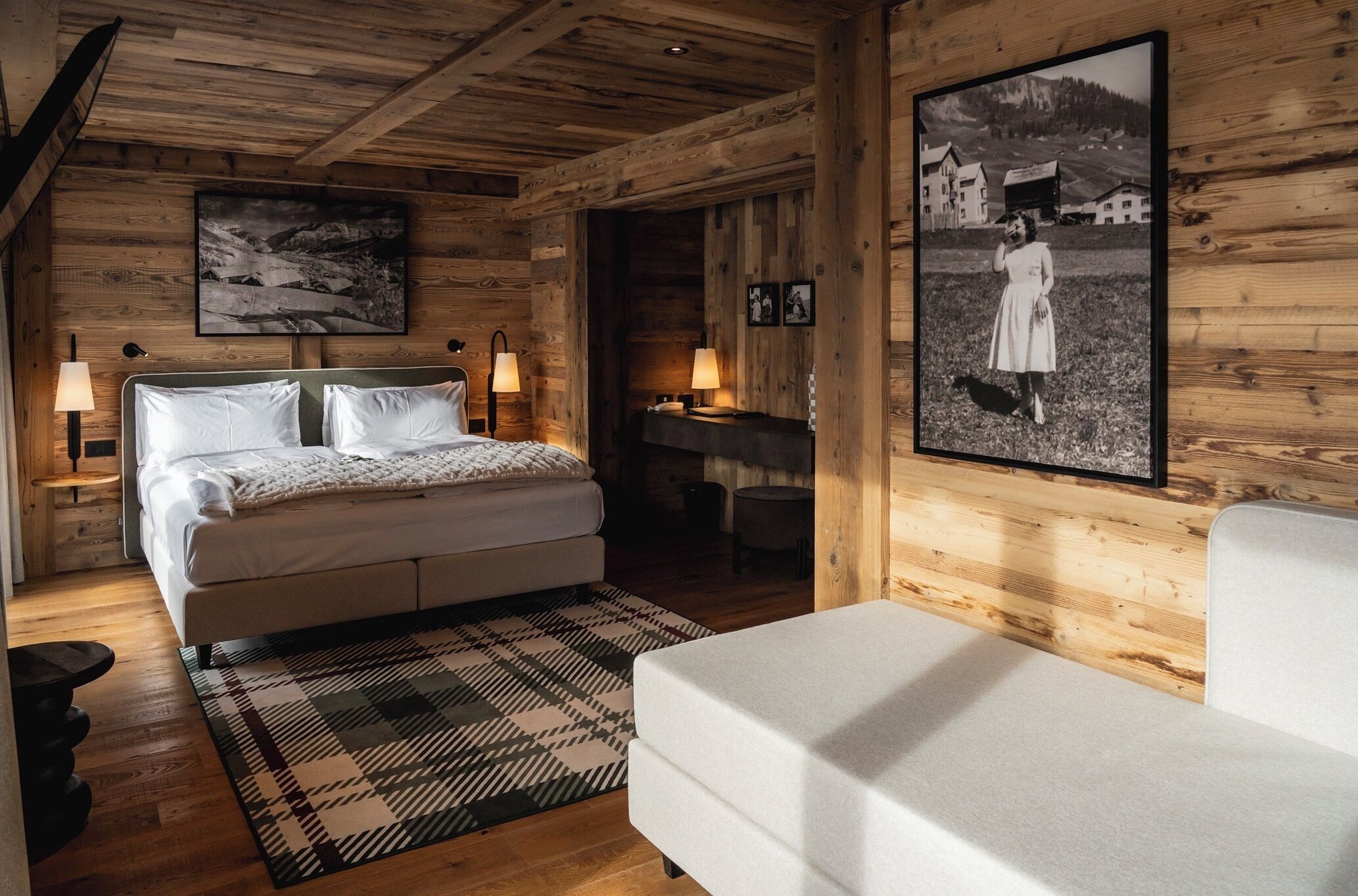 A wooden cabin bedroom with a bed, two bedside lamps, a patterned rug, a white sofa, and a framed photograph on the wall.