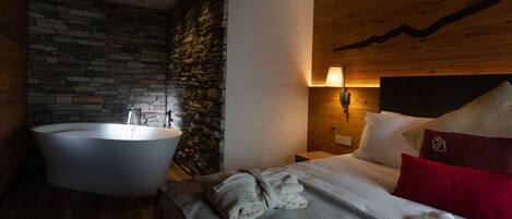 A modern bedroom with a stone wall, a large bathtub, a wooden headboard, and a neatly made bed with pillows.