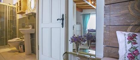 Un baño con cabina de ducha de vidrio, un lavamanos y un inodoro. Hay un espejo sobre el lavamanos y una puerta con pomo. Un jarrón con flores está sobre una mesita junto a la puerta.