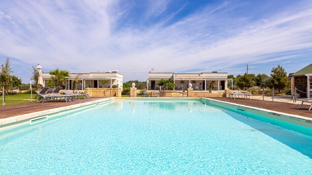 Una piscina con sillas de descanso y sombrillas, rodeada de edificios y vegetación.
