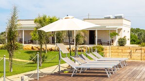 Un área junto a la piscina con sillones de descanso y un toldo, una pared de piedra y un edificio al fondo.