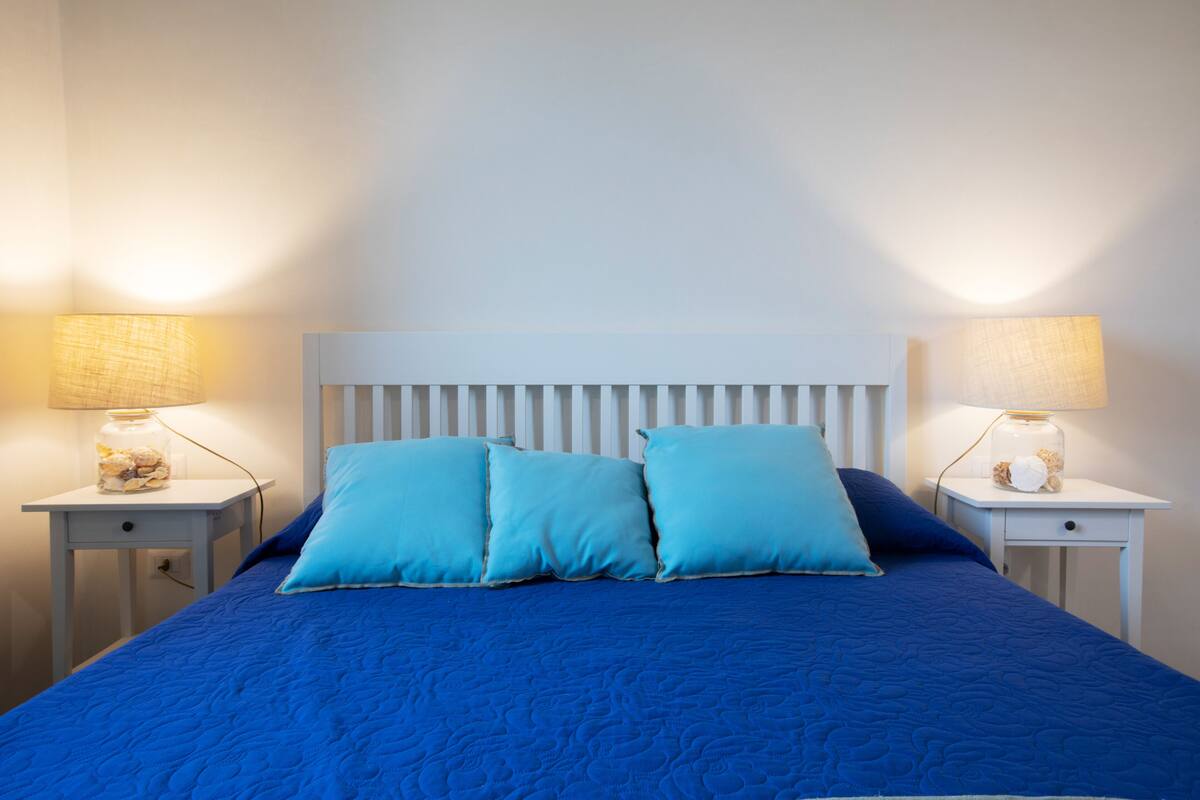 Una cama con colcha azul y dos almohadas, flanqueada por dos mesitas de noche con lámparas y un cabecero blanco.
