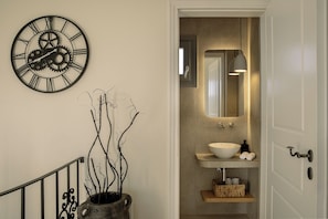 A hallway with a wall-mounted clock, a potted plant, a mirror, and a bathroom with a sink and towel rack.