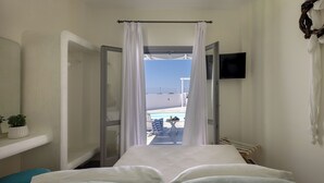 Quarto de hotel com cama, televisão, uma pequena mesa com bebida e vista para a piscina e céu.