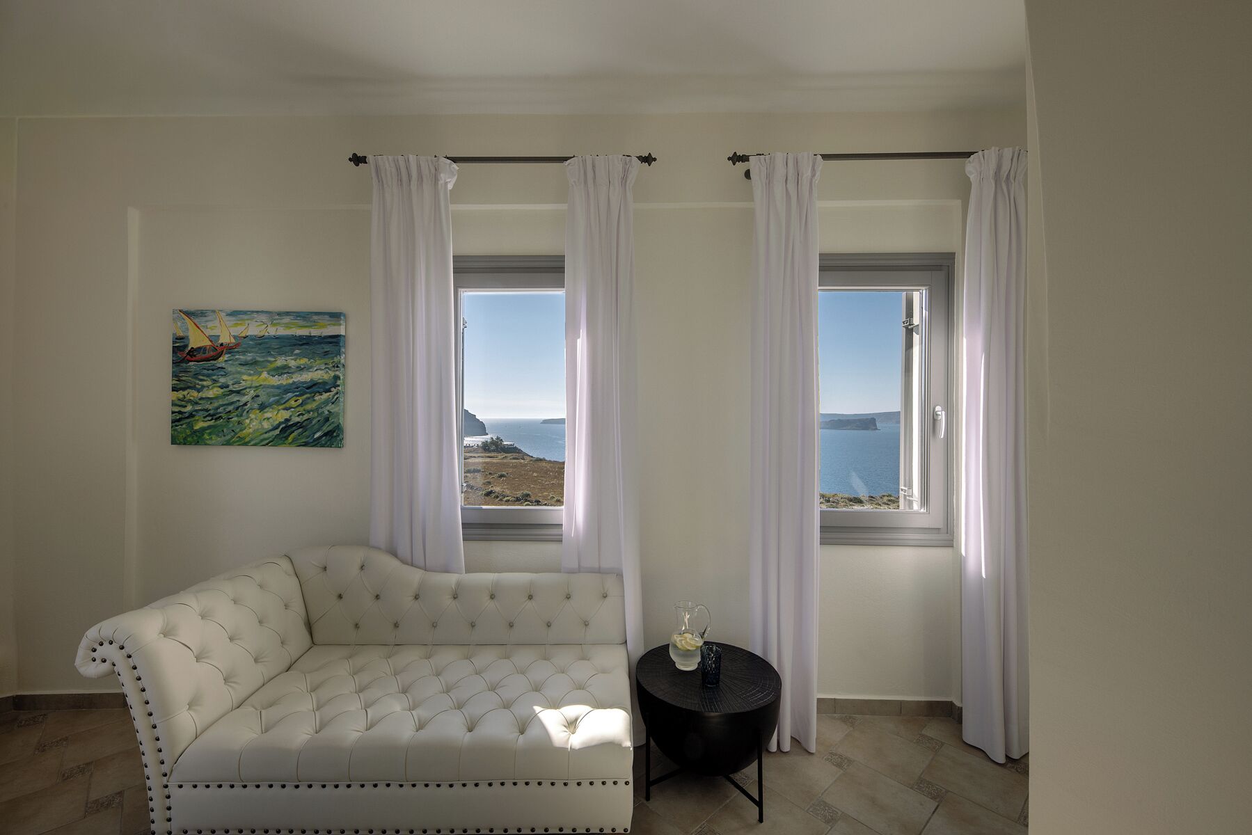 Una stanza con un divano bianco, un tavolino rotondo con un boccale e un bicchiere, e un quadro raffigurante una barca a vela appeso al muro.
