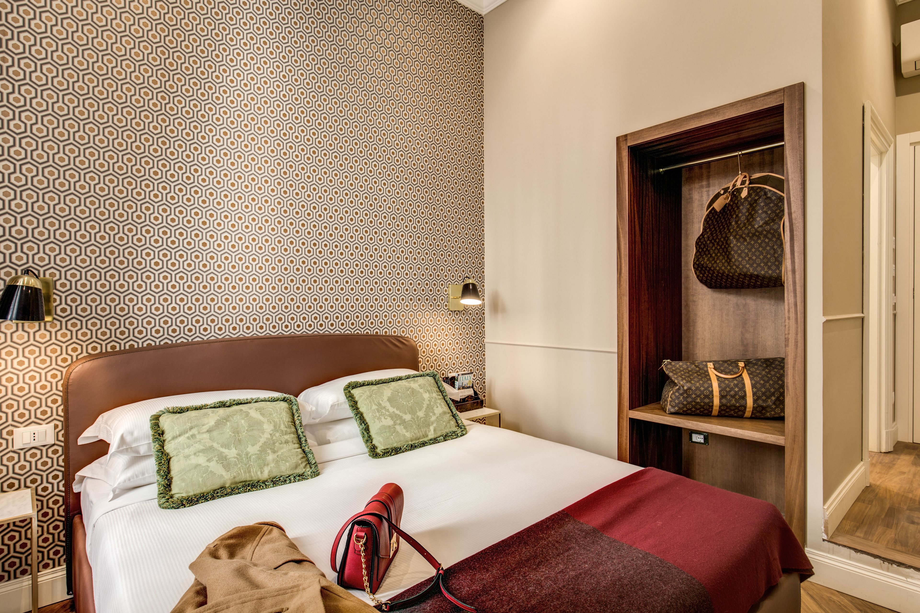 A hotel room with a patterned wallpaper, a bed with green and white bedding, a red throw blanket, a brown robe, and a handbag hanging on a shelf.