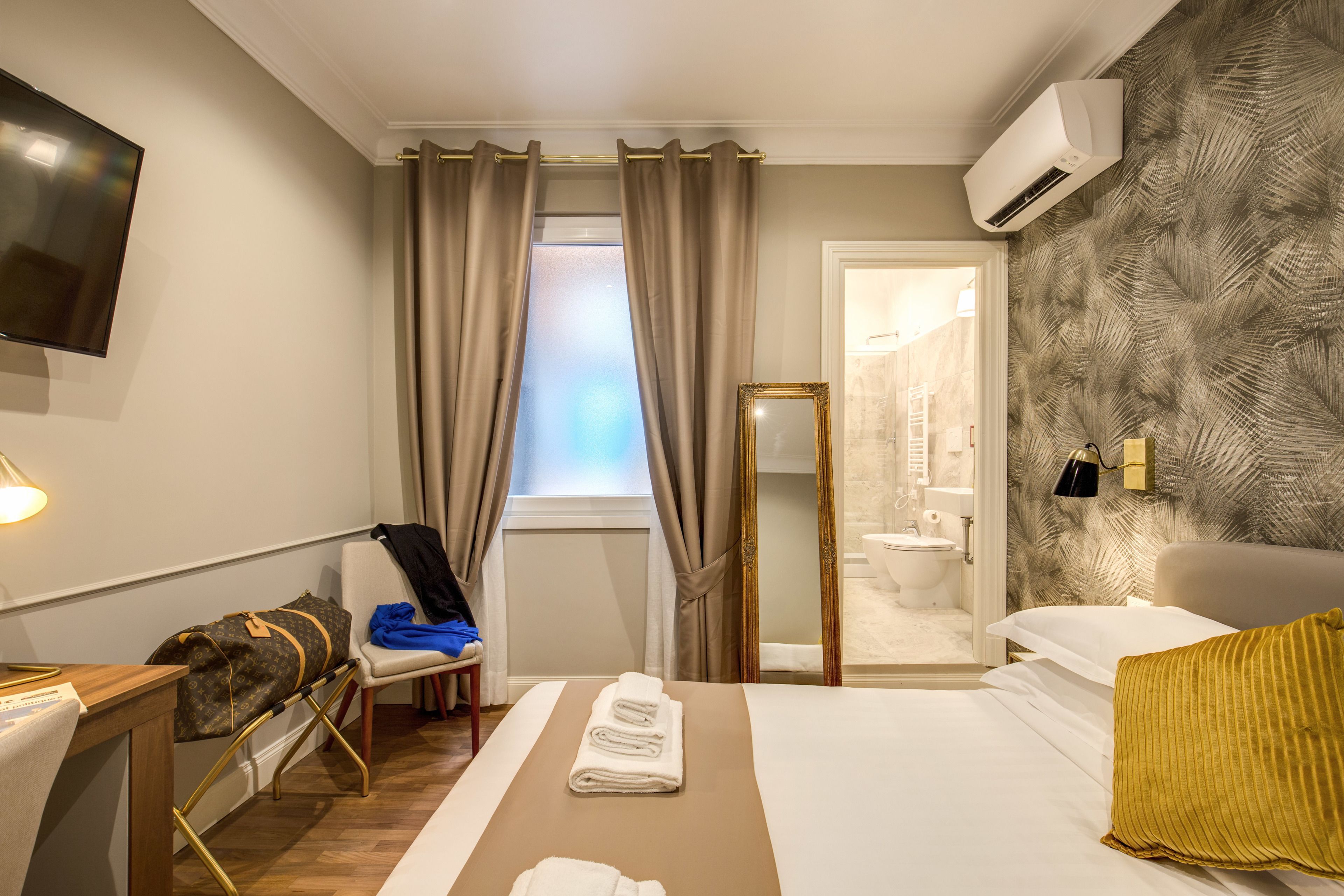 A hotel room with a bed, a desk, a chair, a bathroom visible through an open door, and a mounted TV.