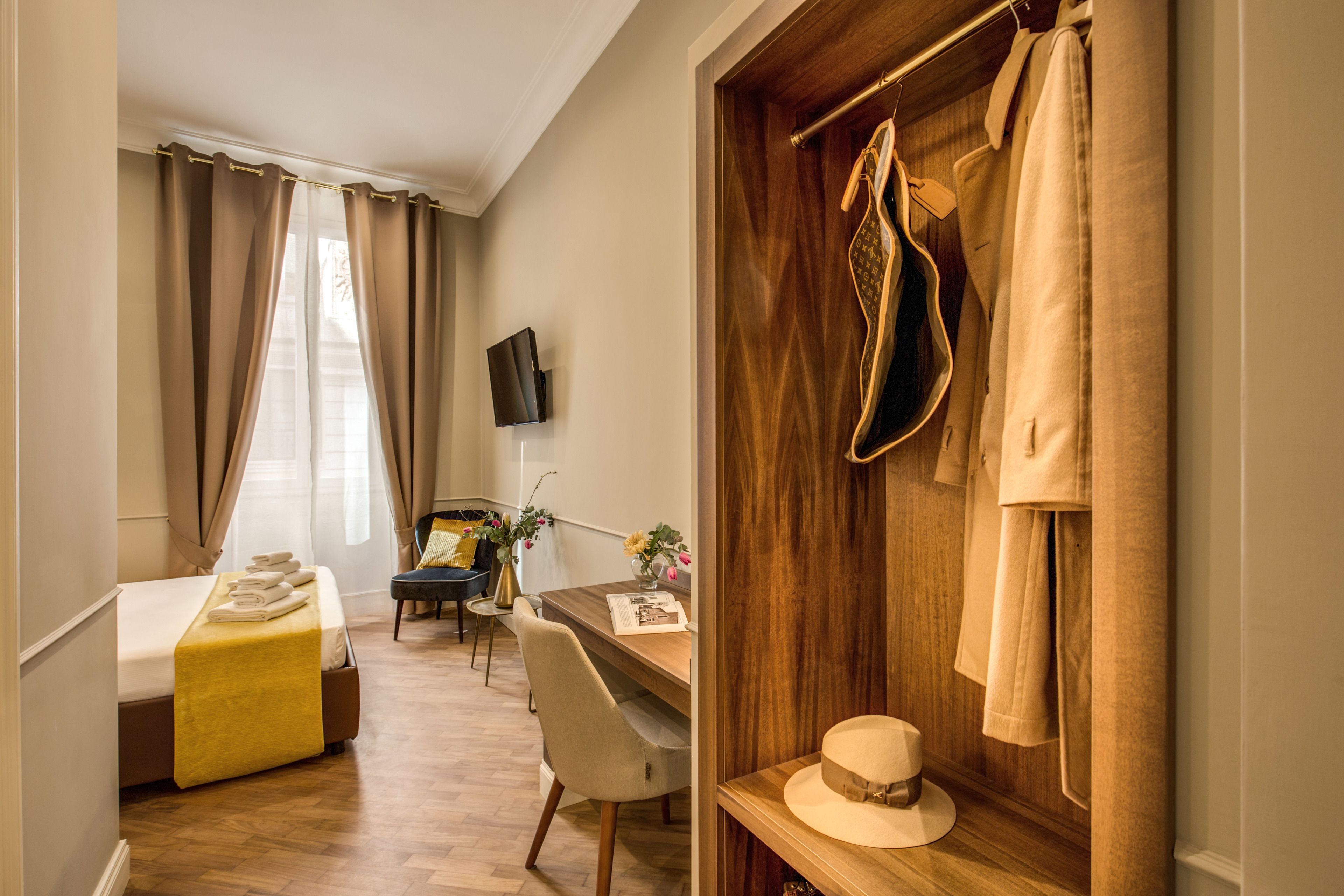 A hotel room with a bed, a desk, a chair, a television, and a wardrobe with hanging clothes.