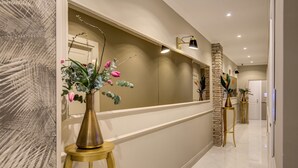 Een gang met een beige muur, een accentmuur van steen, en een witte deur aan het einde. Drie gouden lampen op voetstukken en een bloemenvaas op een gouden standaard.