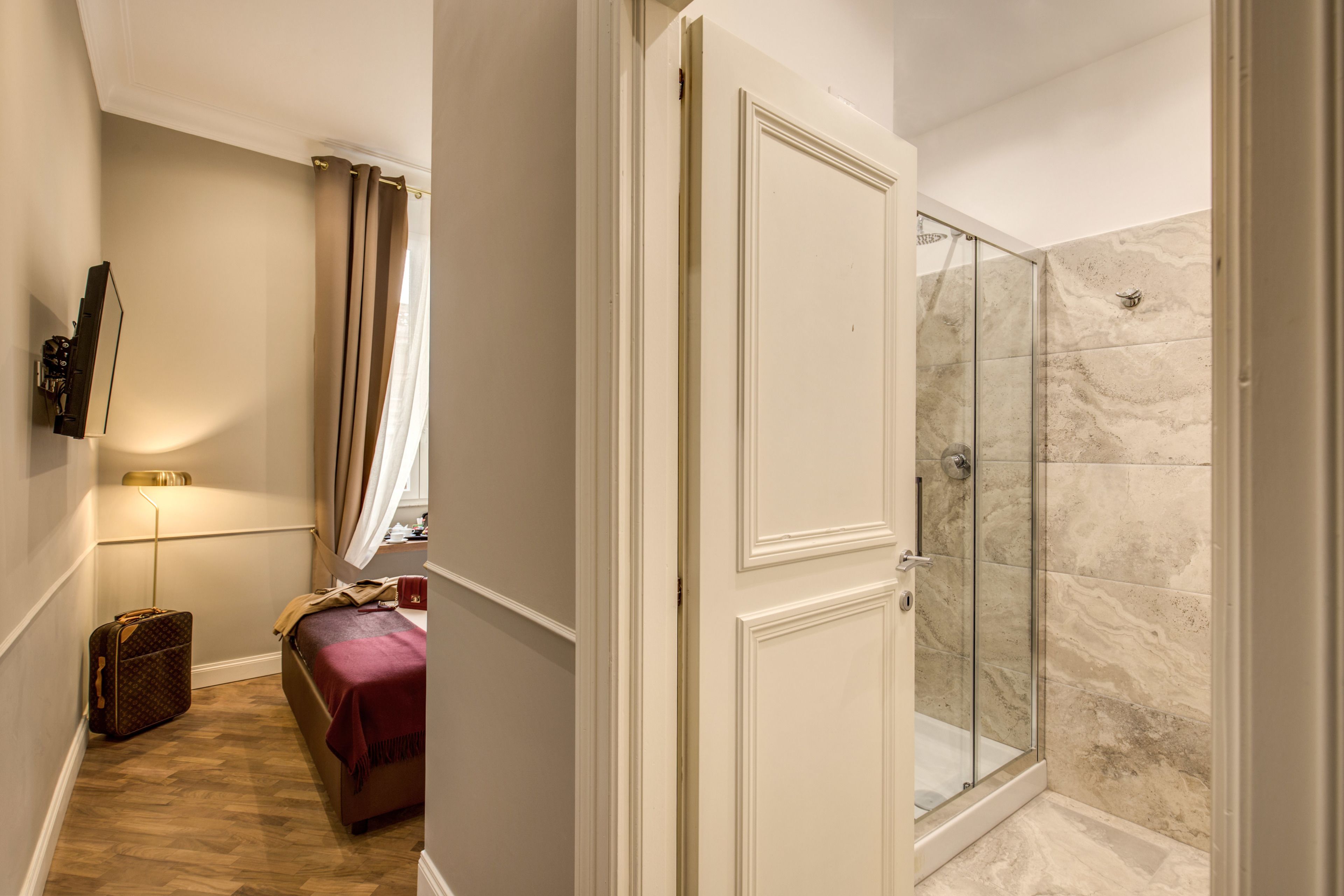 A hotel room with a bed, a suitcase, a glass shower, and a door leading to another room.