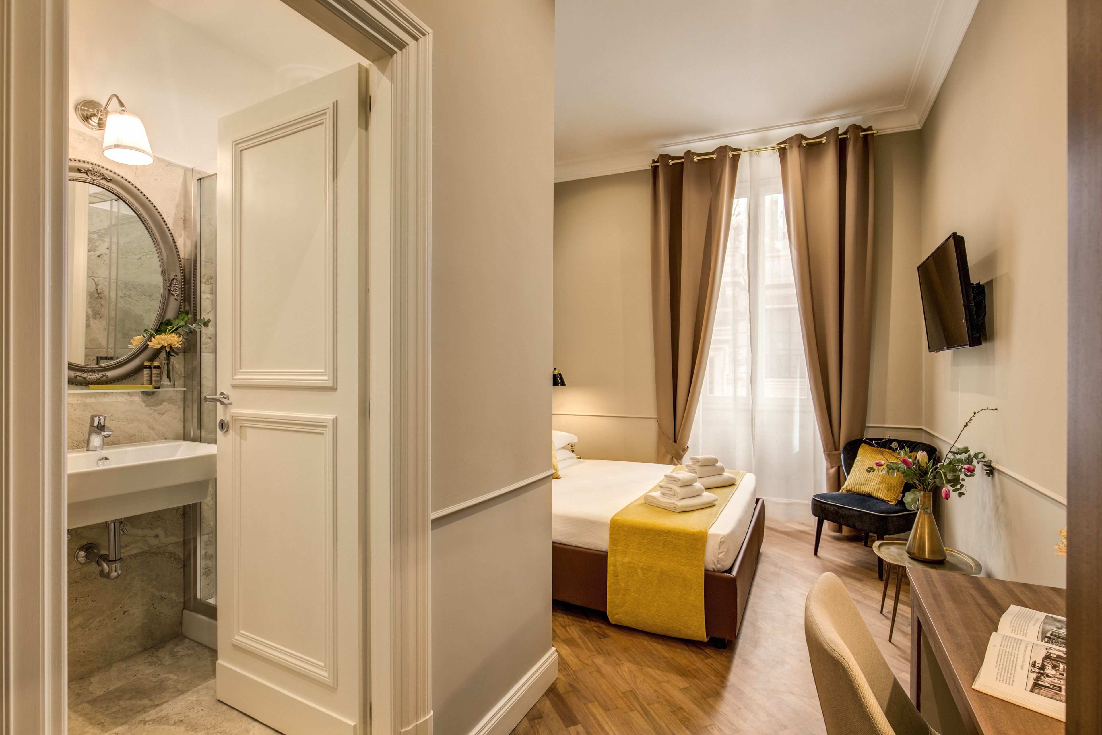 Una habitación de hotel con baño, una cama con colcha amarilla, una silla, una mesita con un florero y un televisor de pantalla plana montado en la pared.