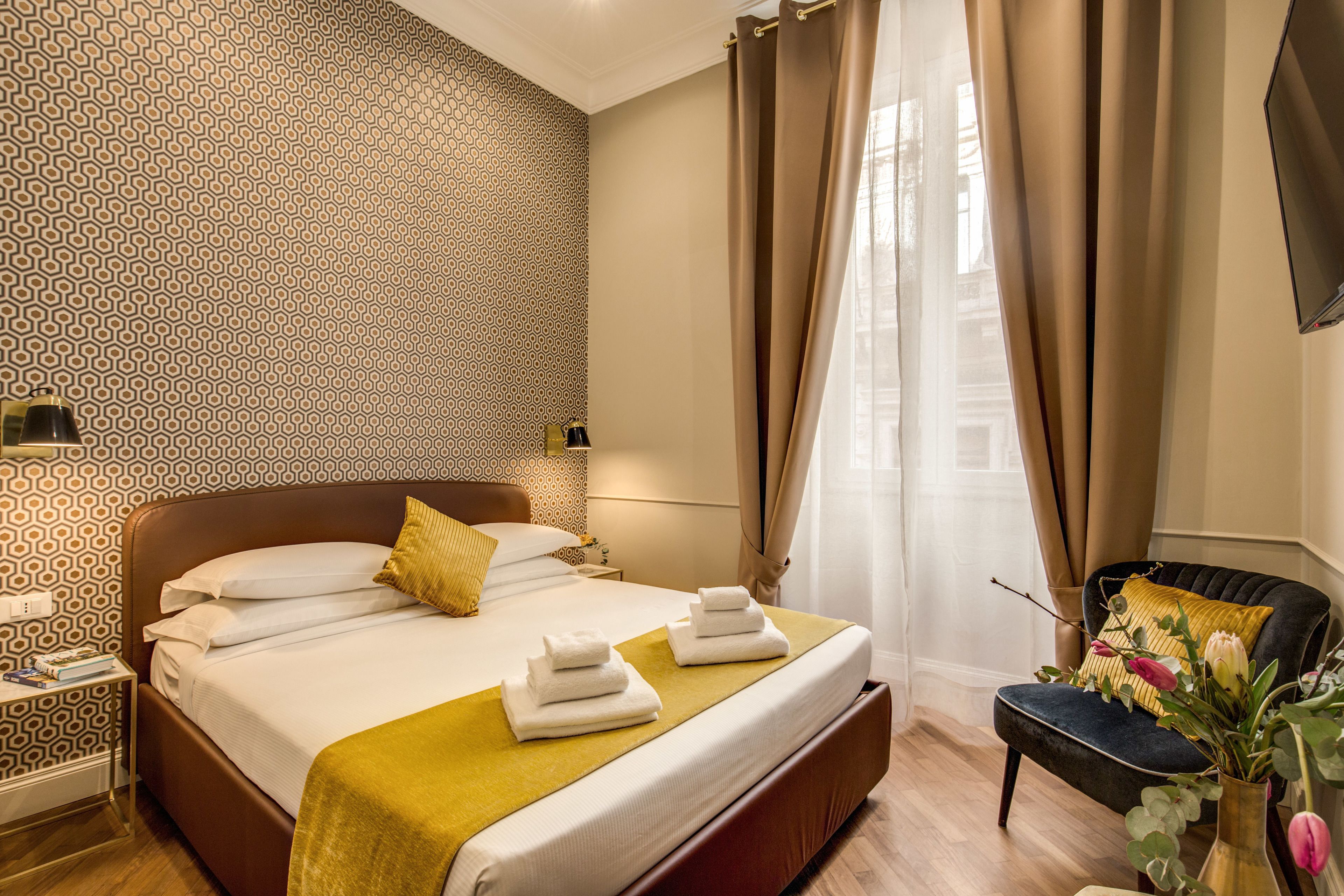 A hotel room with a patterned wallpaper, a bed with white linens and a yellow throw, a nightstand with a lamp, and a chair with a vase of flowers.