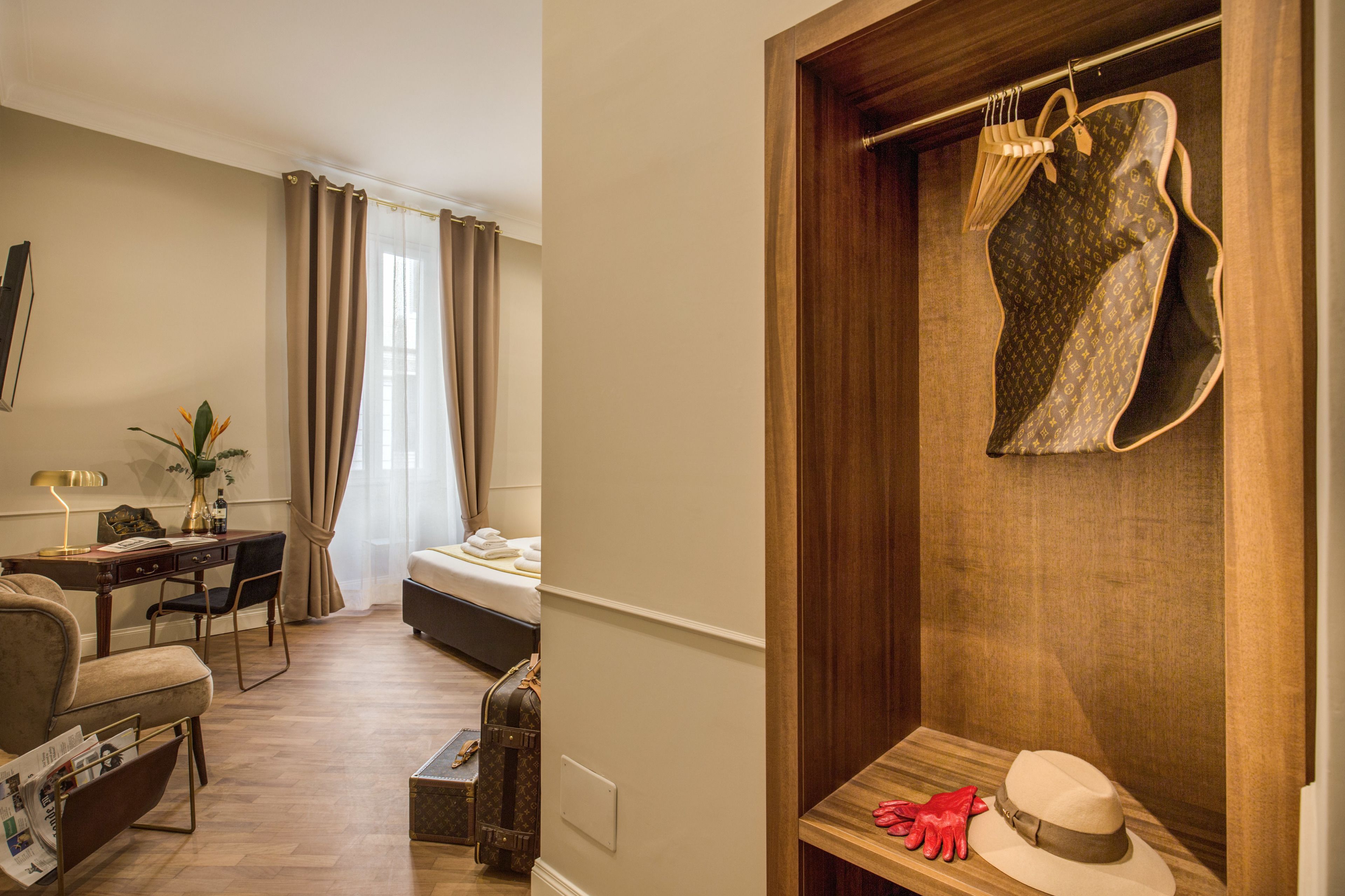 Une chambre d’hôtel avec un bureau en bois, un lit, un coin salon avec un fauteuil et une armoire contenant des vêtements suspendus et un chapeau.