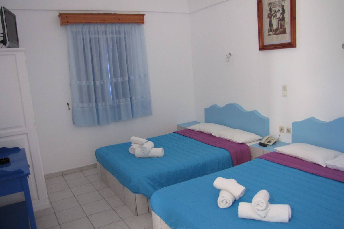 Habitación de hotel con dos camas, cabecera de madera, mesita de noche pequeña con un teléfono y ventana con cortina azul.