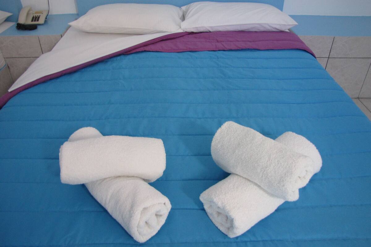 Una cama con una colcha azul, dos toallas blancas enrolladas y una almohada blanca.