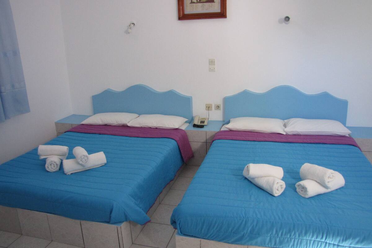 Habitación de hotel con dos camas, cada una con ropa de cama azul y almohadas blancas. Hay toallas sobre las camas y un teléfono en la mesita de noche.