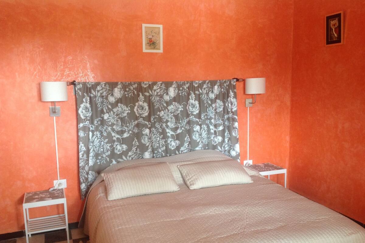 A bedroom with an orange wall, a floral-patterned headboard, two bedside lamps, and a bed with a quilted cover.