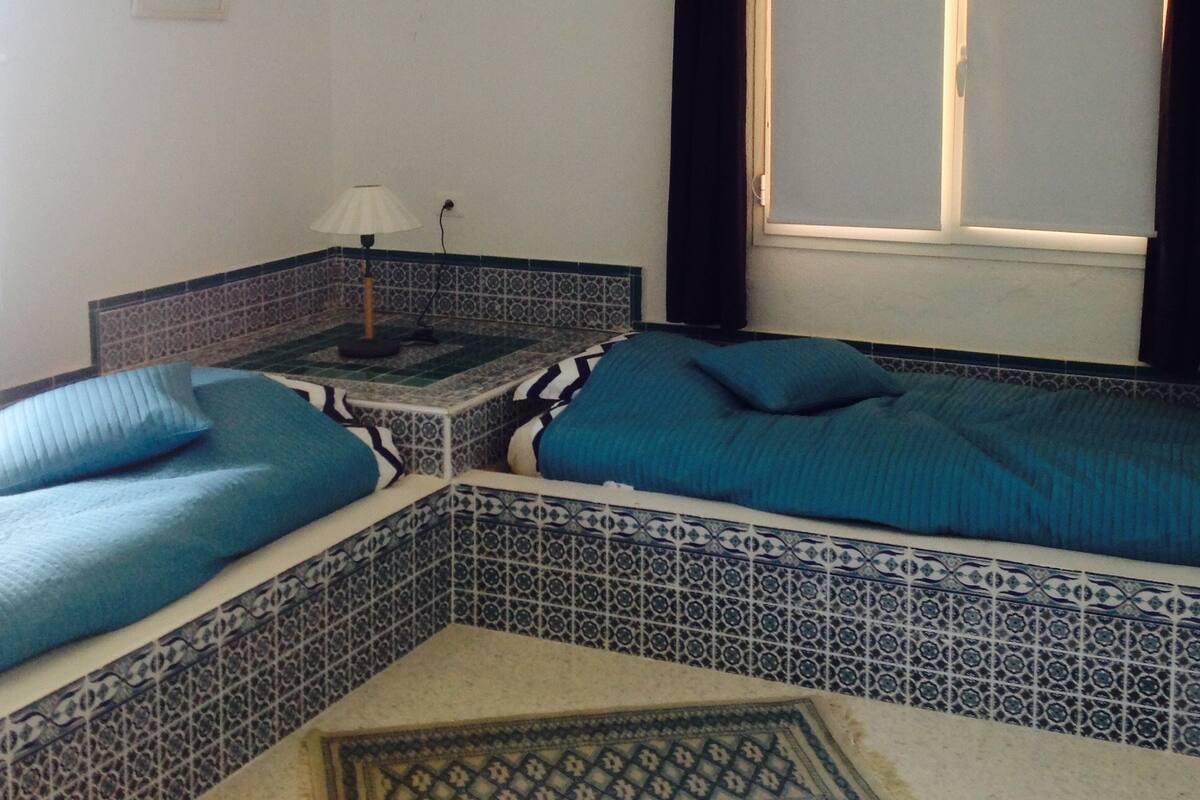 A room with a bed, a nightstand, a lamp, an air conditioner, a window with curtains, and a patterned tile backsplash.