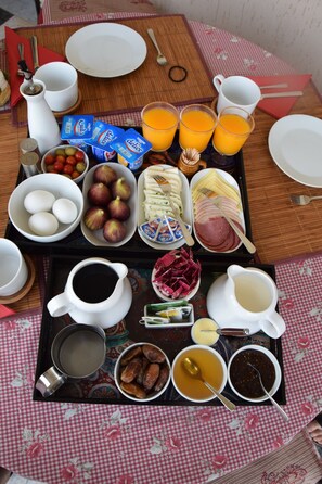 Bandeja de desayuno con huevos, frutas, queso, jamón y bebidas sobre un mantel a cuadros.