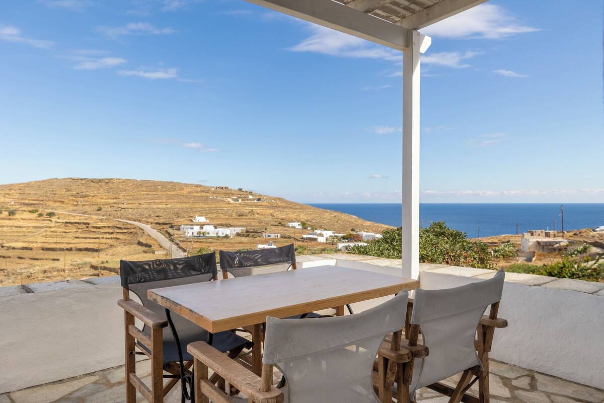 An outdoor patio with a wooden table and chairs, overlooking a coastal landscape with white buildings and a clear blue sky.