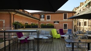 An outdoor patio with a wooden deck, clear glass bar stools, a sofa, and a yellow chair. - MyTALE Creative Academy Hotel (Rome)