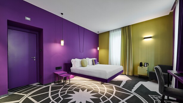 A modern hotel room with a purple wall, a large bed, a desk, and a chair. - MyTALE Creative Academy Hotel (Rome)