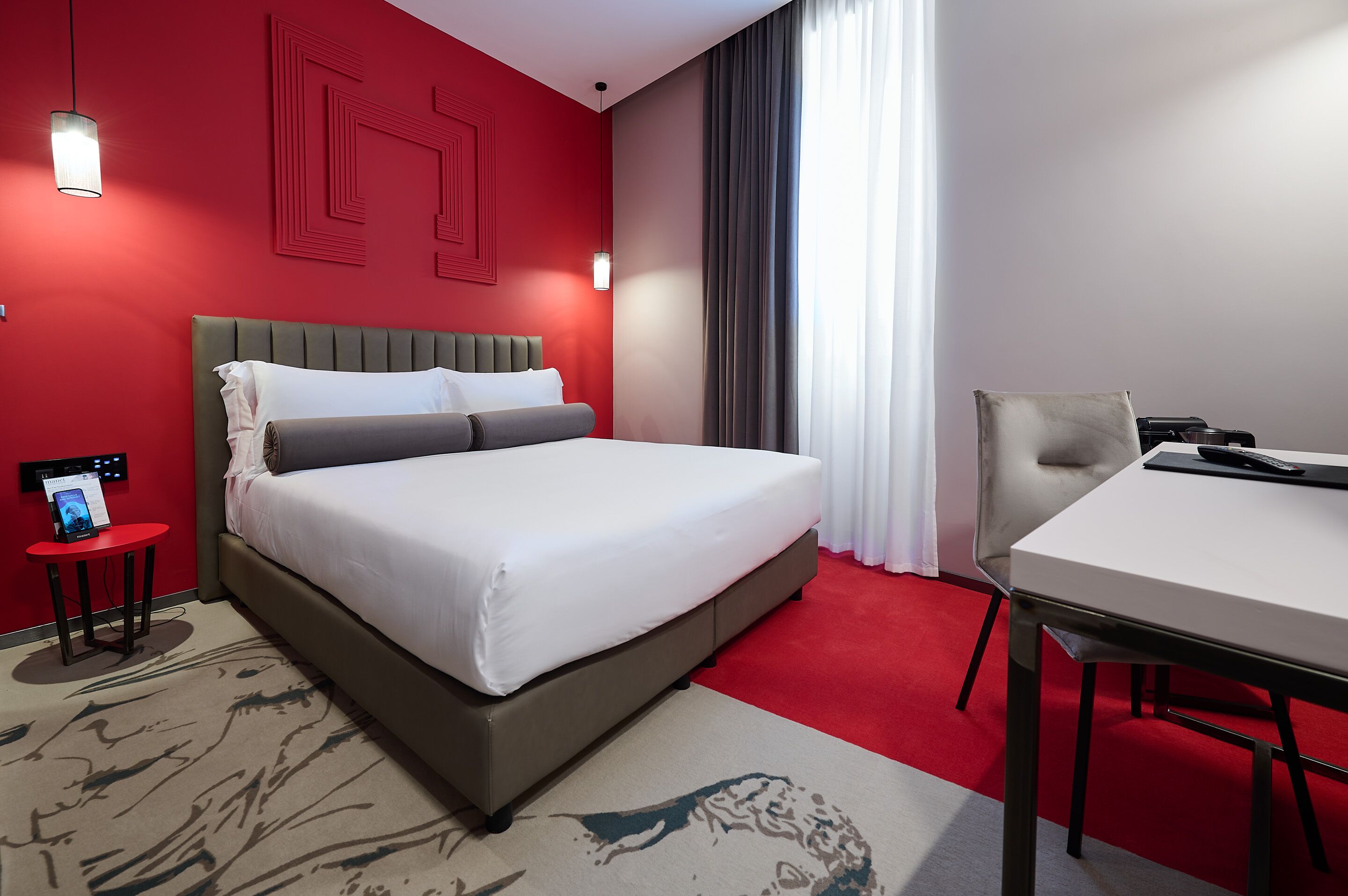 A hotel room with a bed, a desk, a chair, a red wall, and a window with curtains.