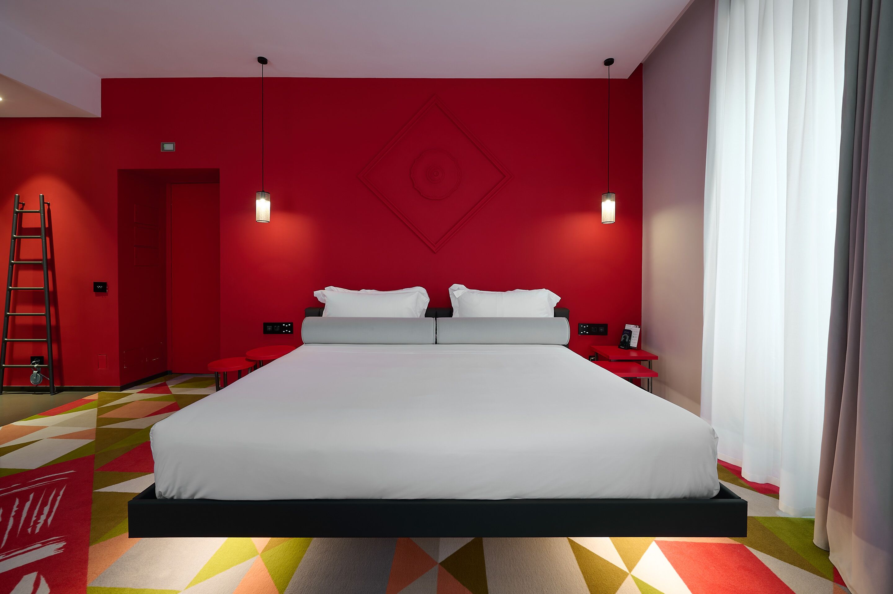 A modern hotel room with a large bed, red walls, and a geometric patterned carpet.
