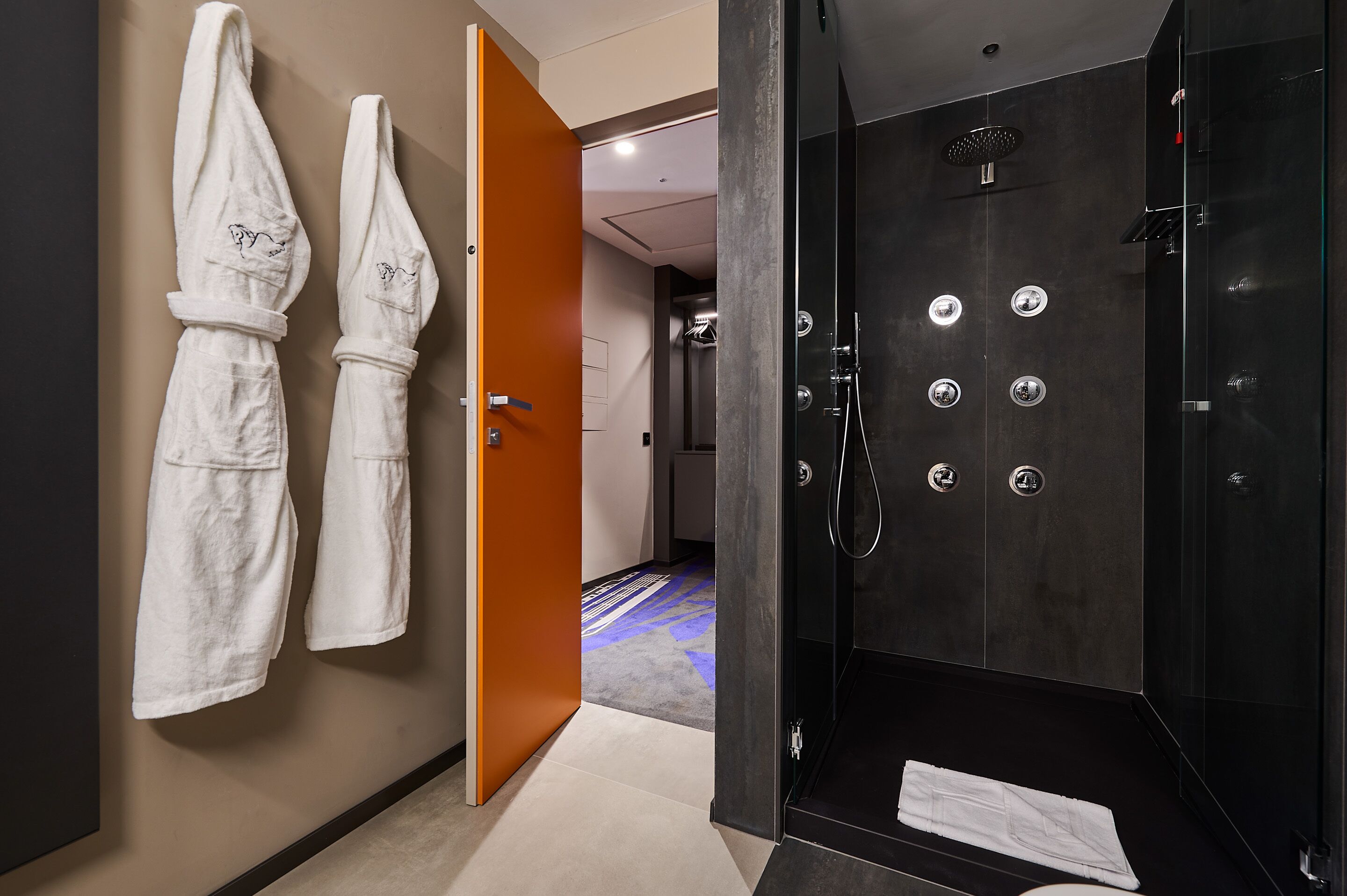 Un bany d'hotel amb una dutxa, dues tovalloles penjades a la paret i una porta visible que condueix a una altra habitació.