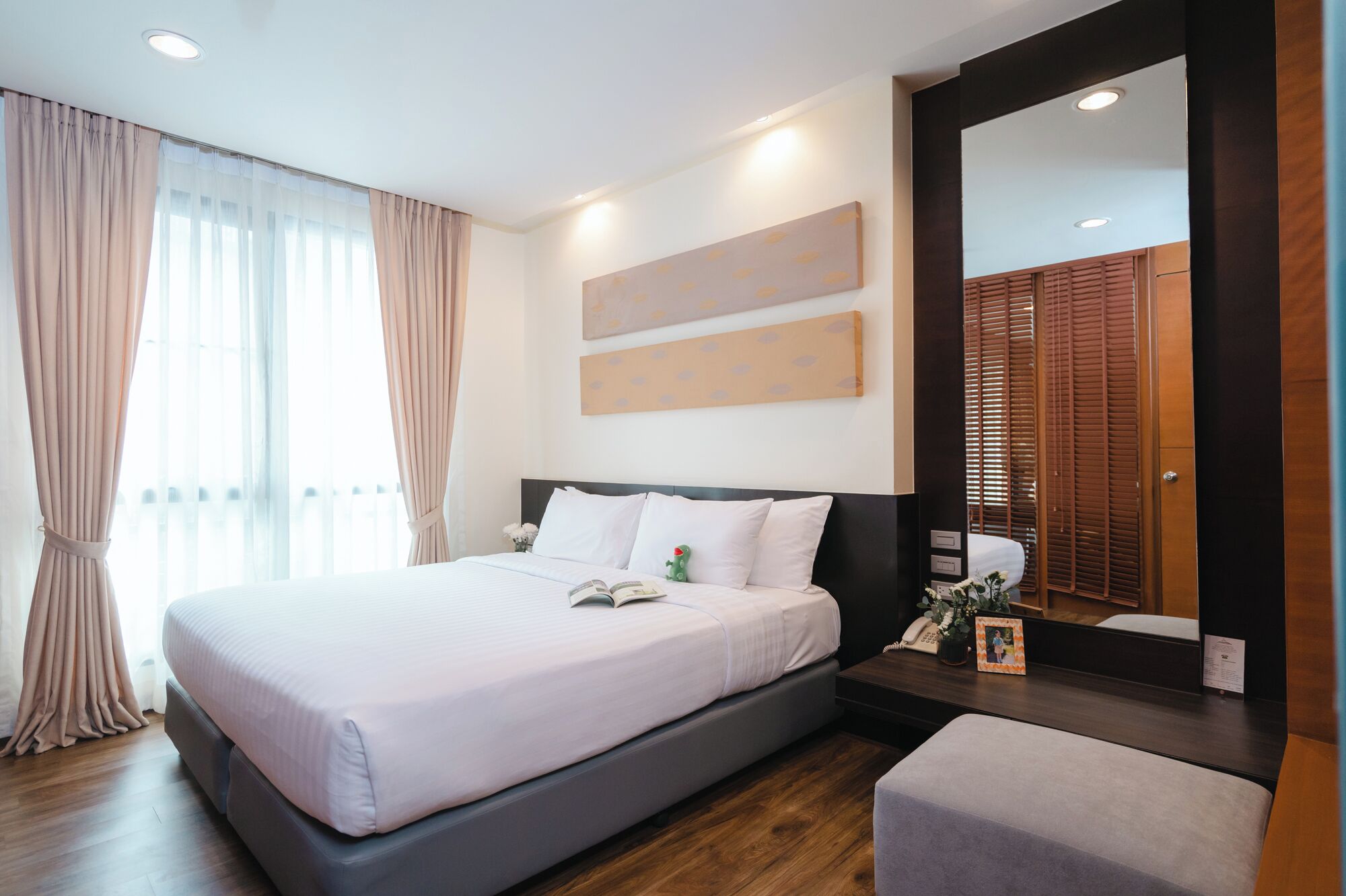 Quarto de hotel moderno com uma cama grande, uma mesa de cabeceira com abajur, um espelho e vista para uma porta de madeira.