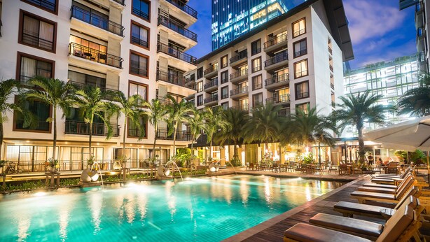 Zona de piscina con varias tumbonas, palmeras y un edificio bien iluminado al fondo.