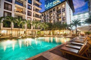 Zona de piscina con varias tumbonas, palmeras y un edificio bien iluminado al fondo.