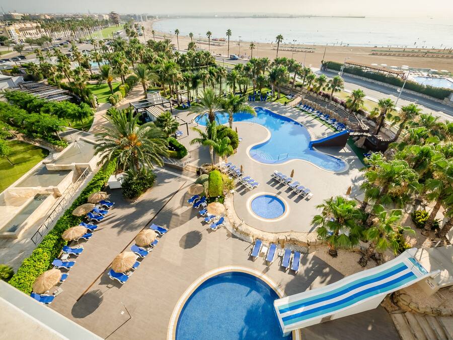 A resort with a large pool, lounge chairs, palm trees, and a beach view.
