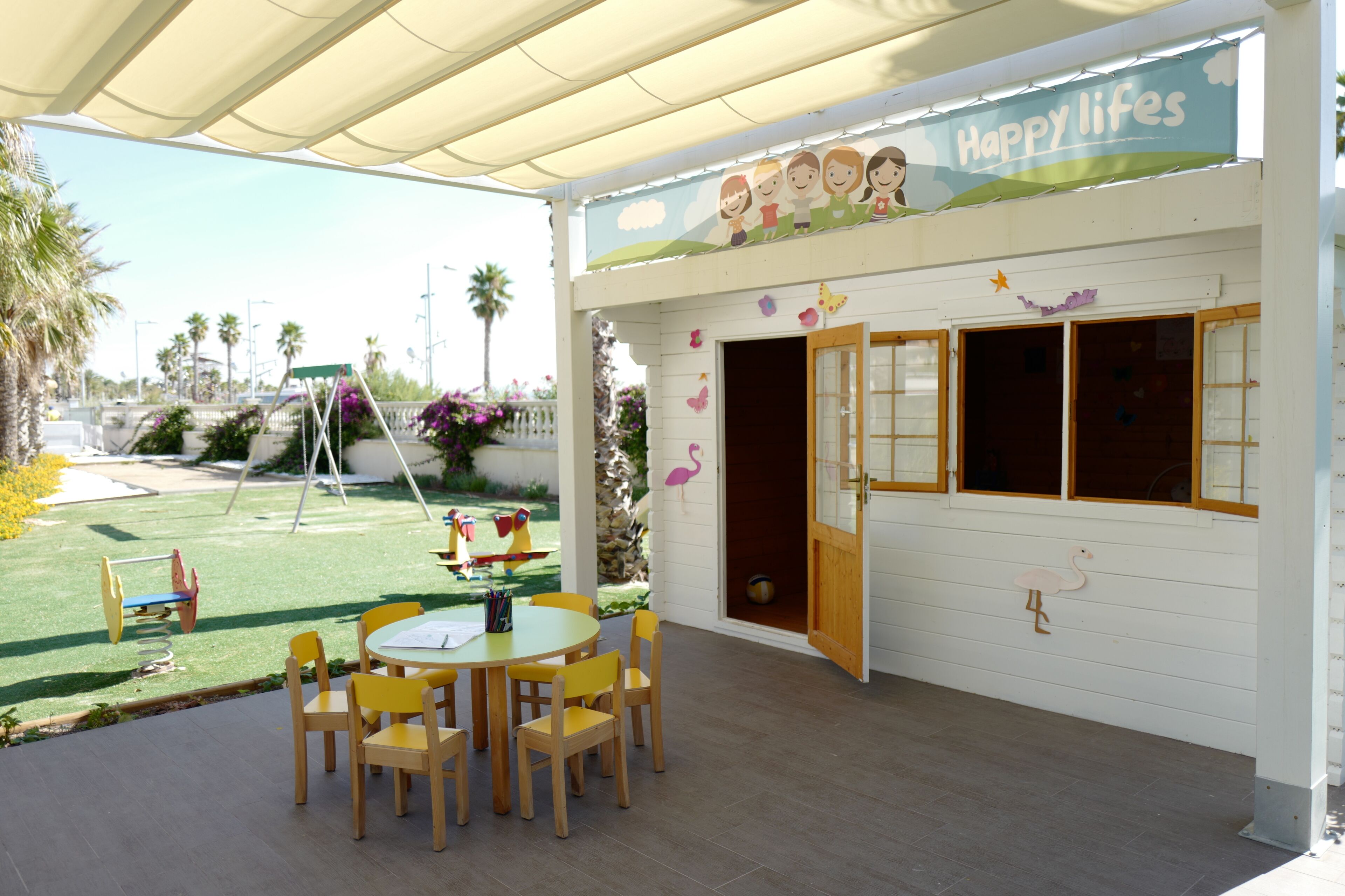 Ein Kinderspielbereich mit Tisch und Stühlen, einer Schaukel und einem Garten mit Blumen und Palmen.