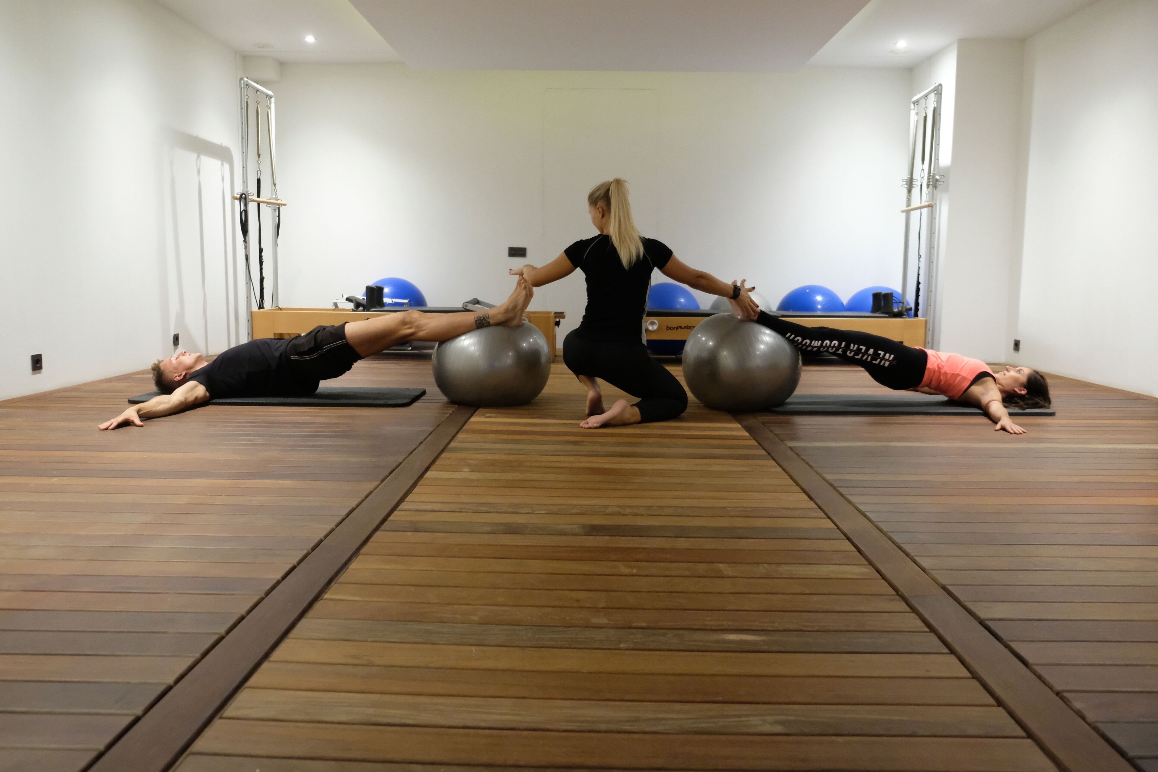 A Pilates class in a room with wooden flooring, exercise balls, and Pilates reformers.