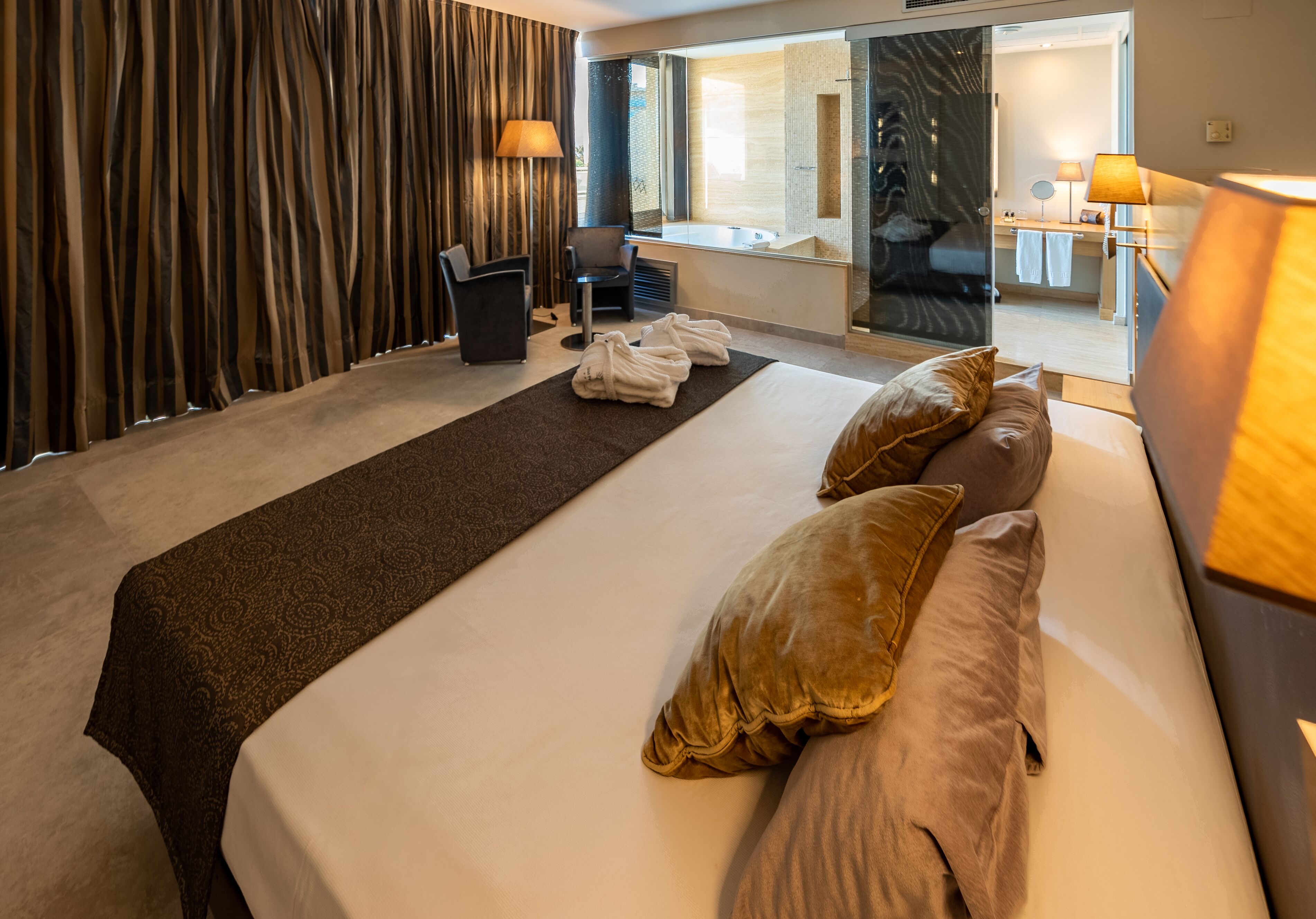 A hotel room with a large bed, a sitting area with a chair, a desk, and a bathroom visible through a glass door.