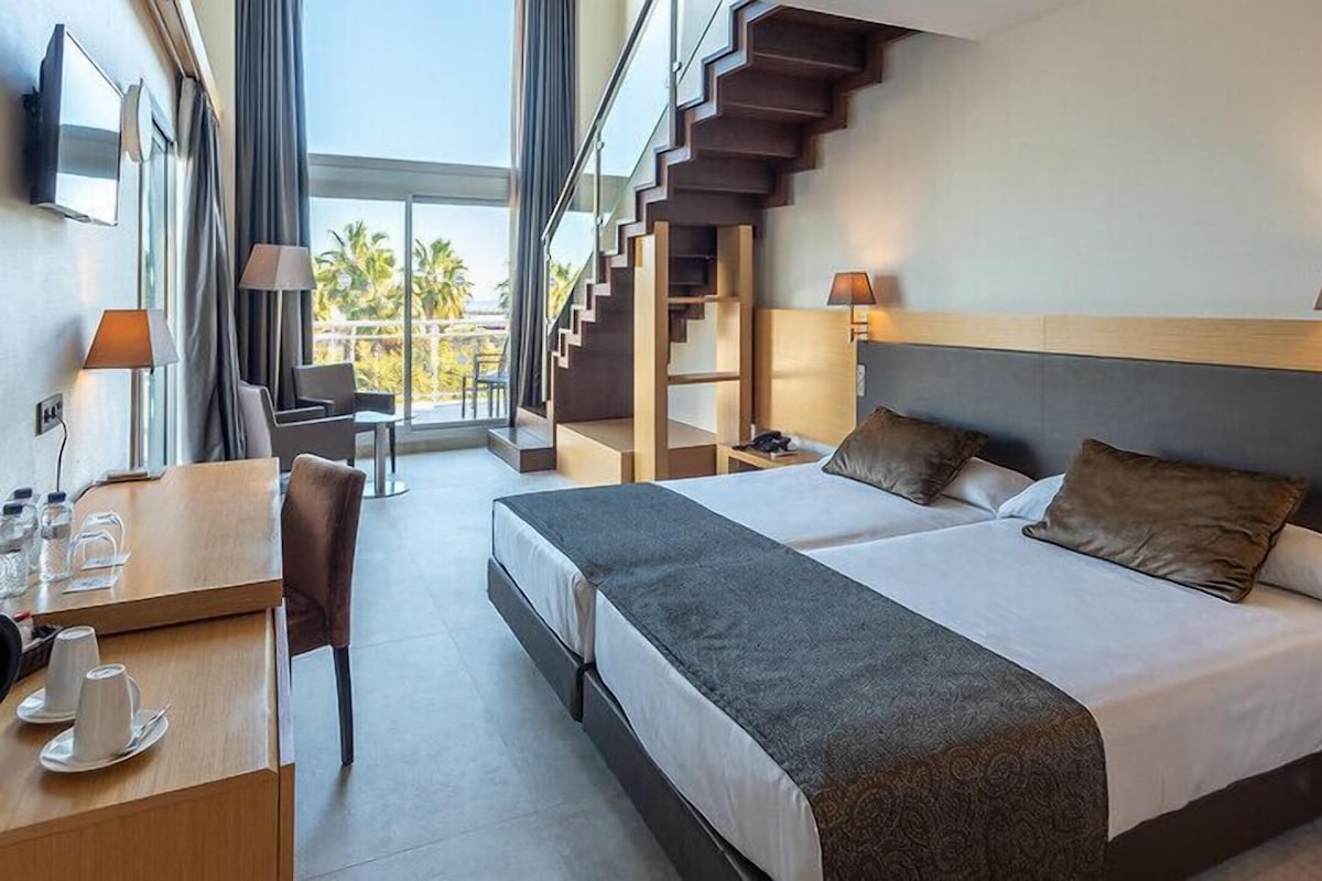 A hotel room with a large bed, a desk with a chair, a lamp, and a view of a staircase leading to a balcony.