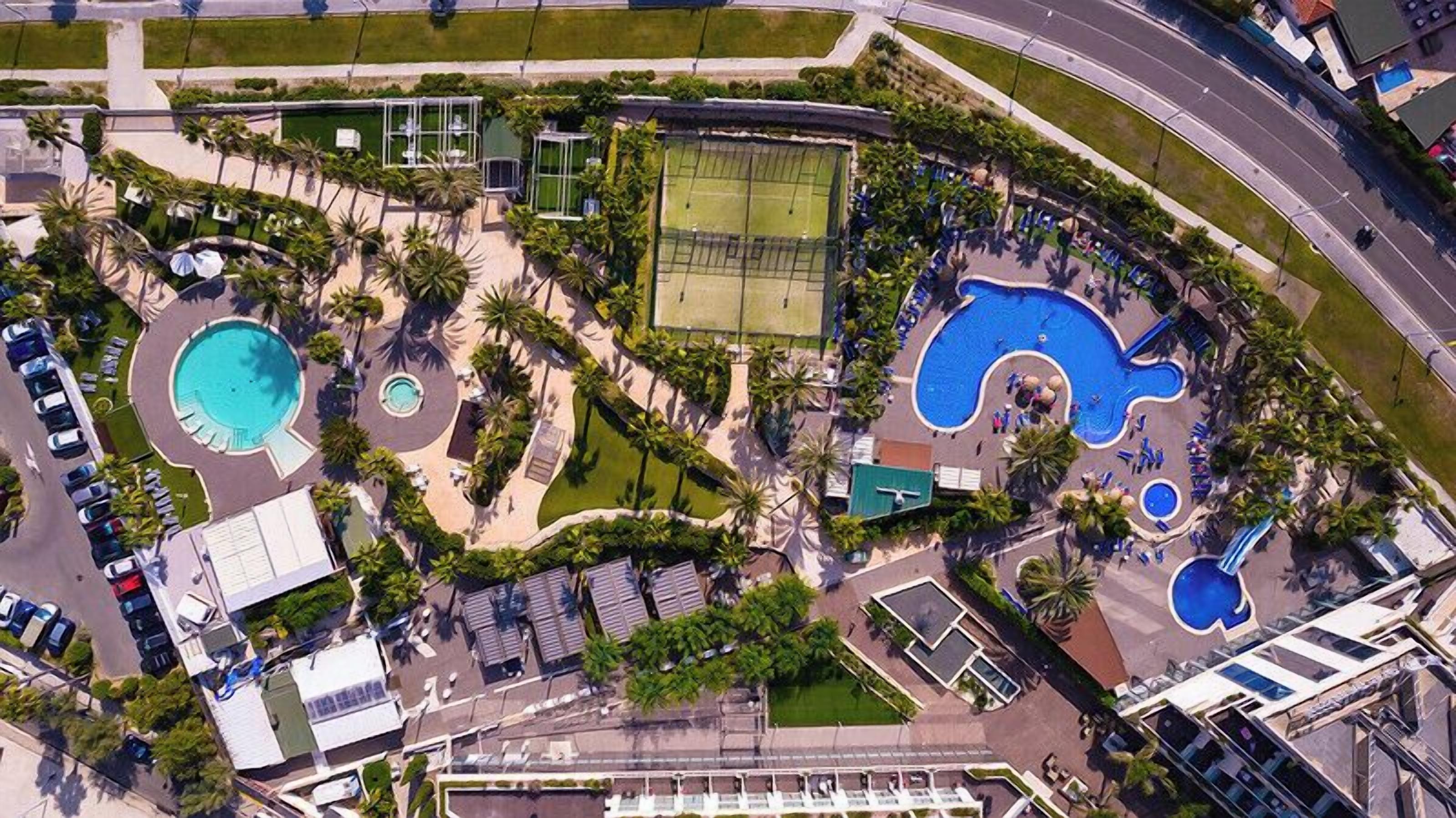 Vista aerea di un resort con diverse piscine, un campo da tennis e un’area parcheggio.