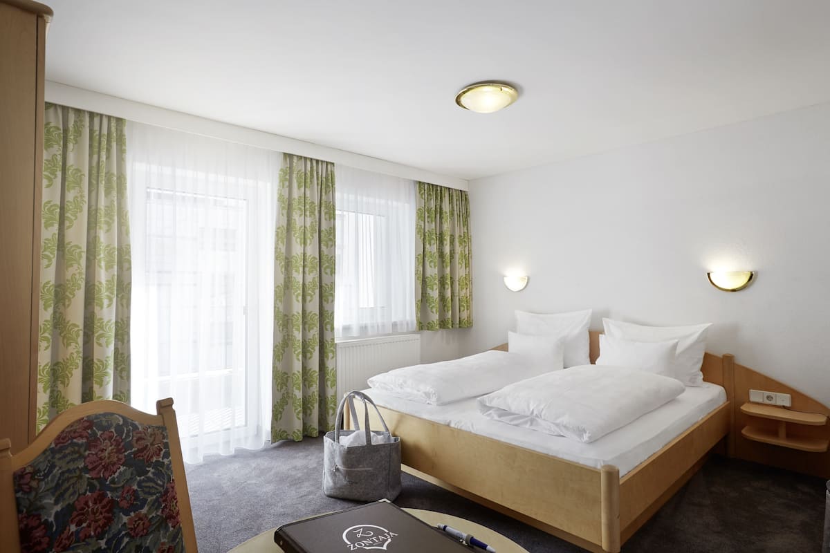 A hotel room with a wooden bed, two bedside tables, a nightstand, a chair, a desk with a laptop, and a window with sheer curtains.