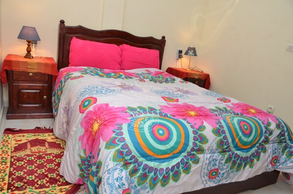 Un lit recouvert d’une couvre-lit aux motifs floraux colorés, deux tables de chevet avec des lampes et un tapis à motifs sur le sol.
