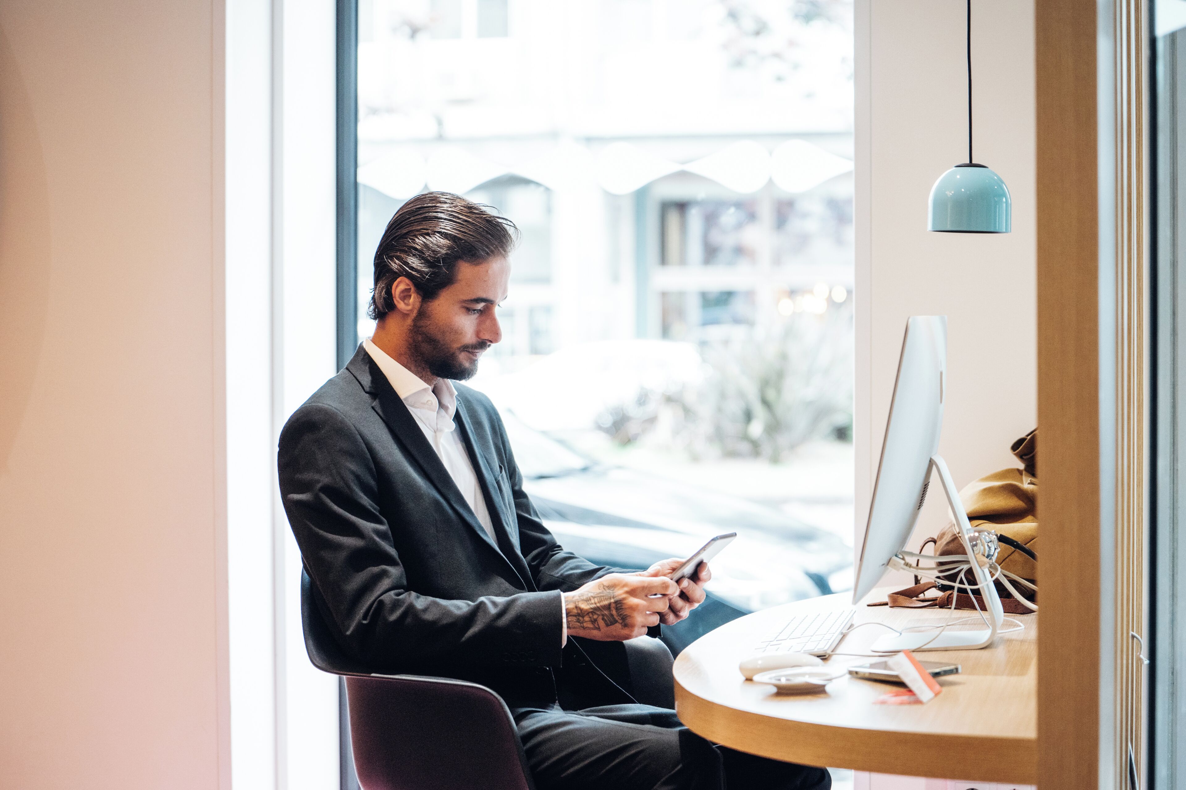 A man in a suit sitting at a desk with a computer and a smartphone.