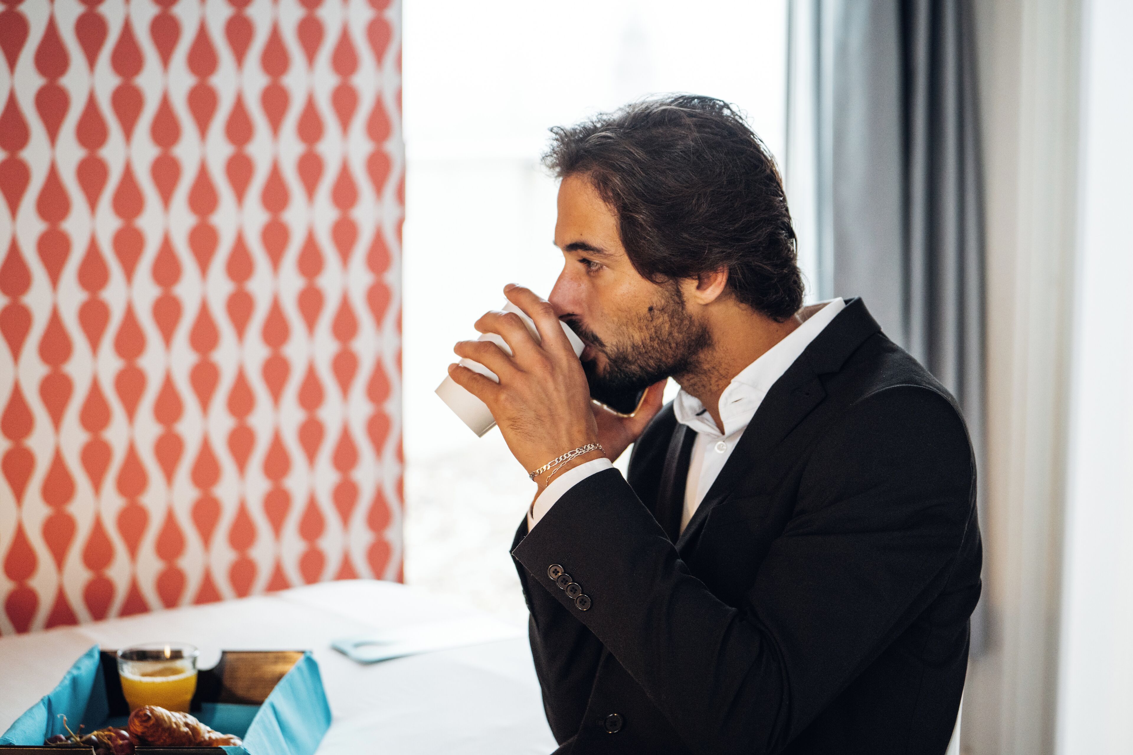 Un uomo in abito nero che beve da una tazza bianca, con un vassoio della colazione contenente croissant e succo d'arancia su un letto in una camera d'albergo con carta da parati decorata.