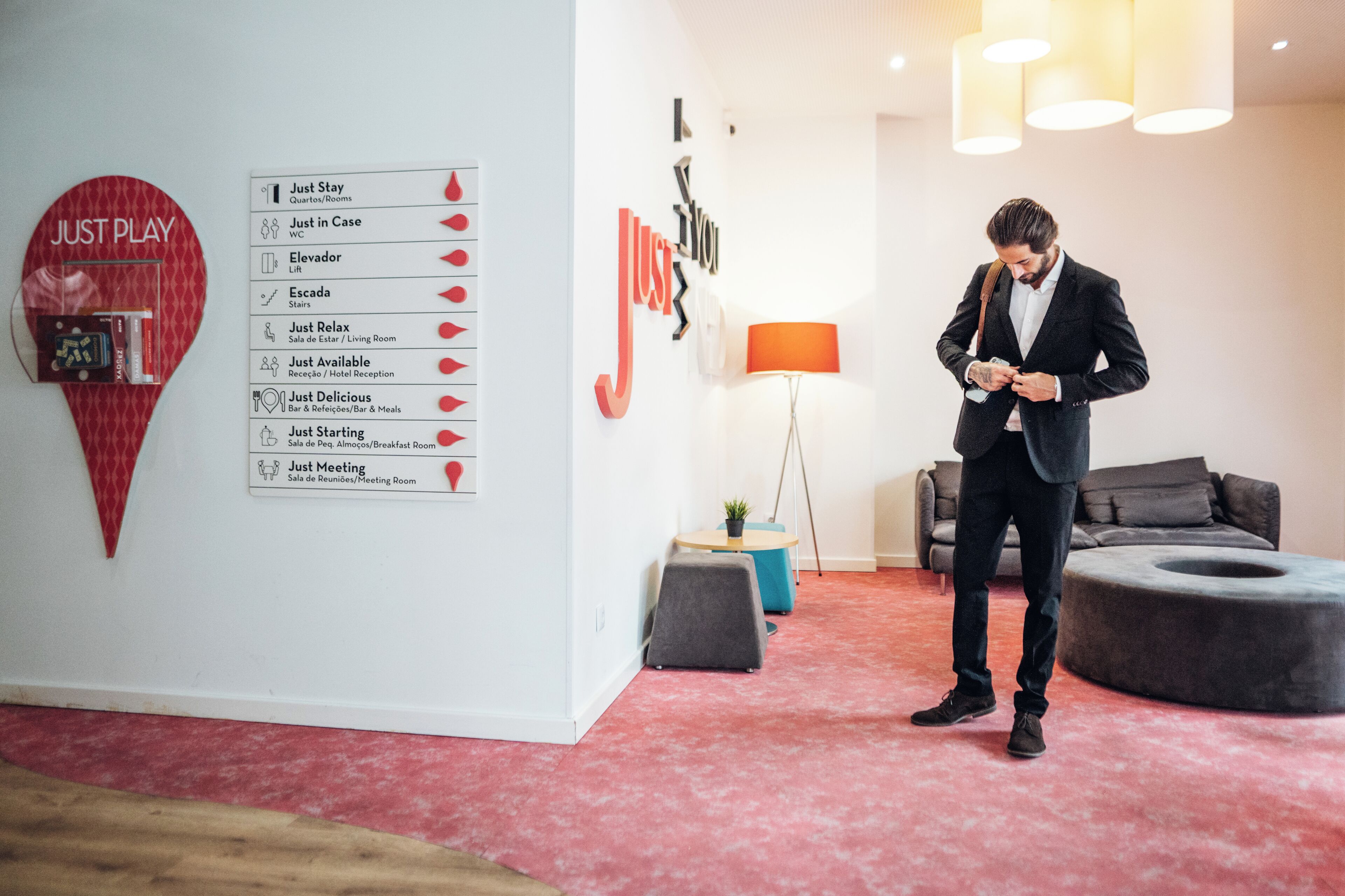 A man in a suit adjusting his tie in a modern hotel room with a red carpet, a red heart-shaped sign, and a wall menu listing amenities like elevators and meeting rooms.