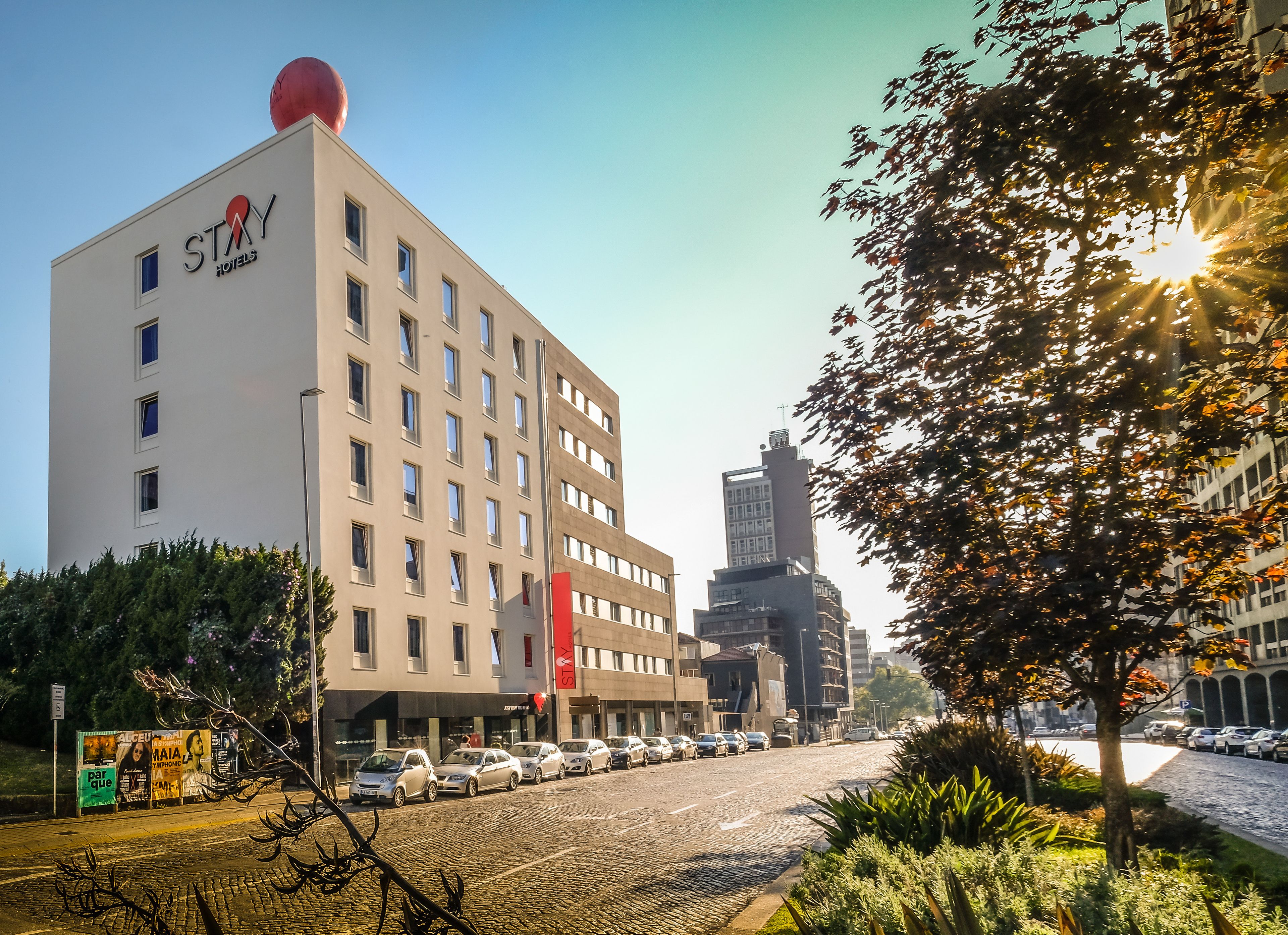A modern hotel with a prominent red logo, multiple floors, and a parking lot with cars.