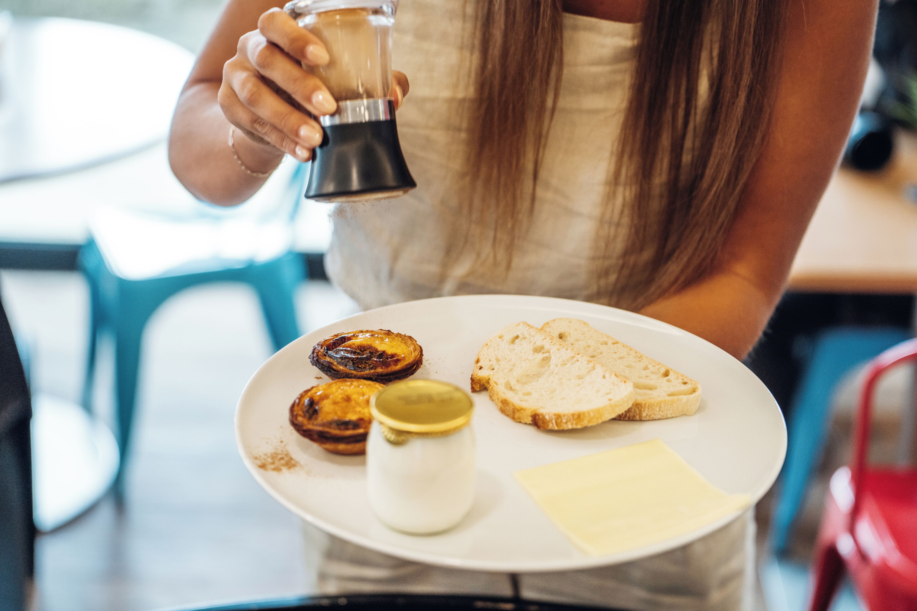 A person holding a coffee grinder, a plate with toast and a small jar, likely containing butter or jam, in a casual dining setting.